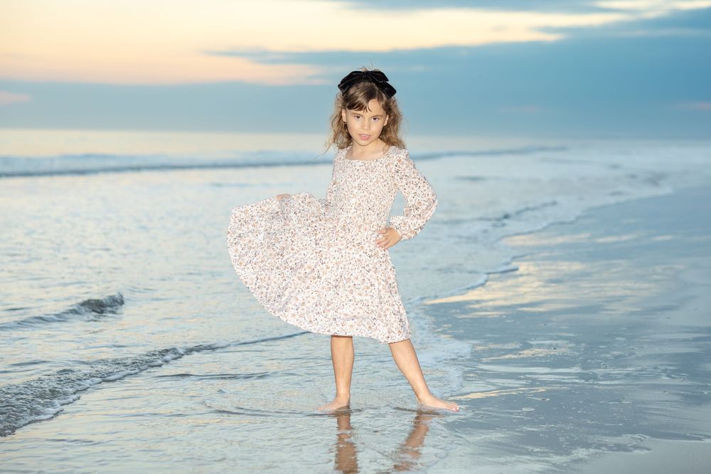 Young girl in a floral dress posing confidently on a beach at sunset with waves in the background.