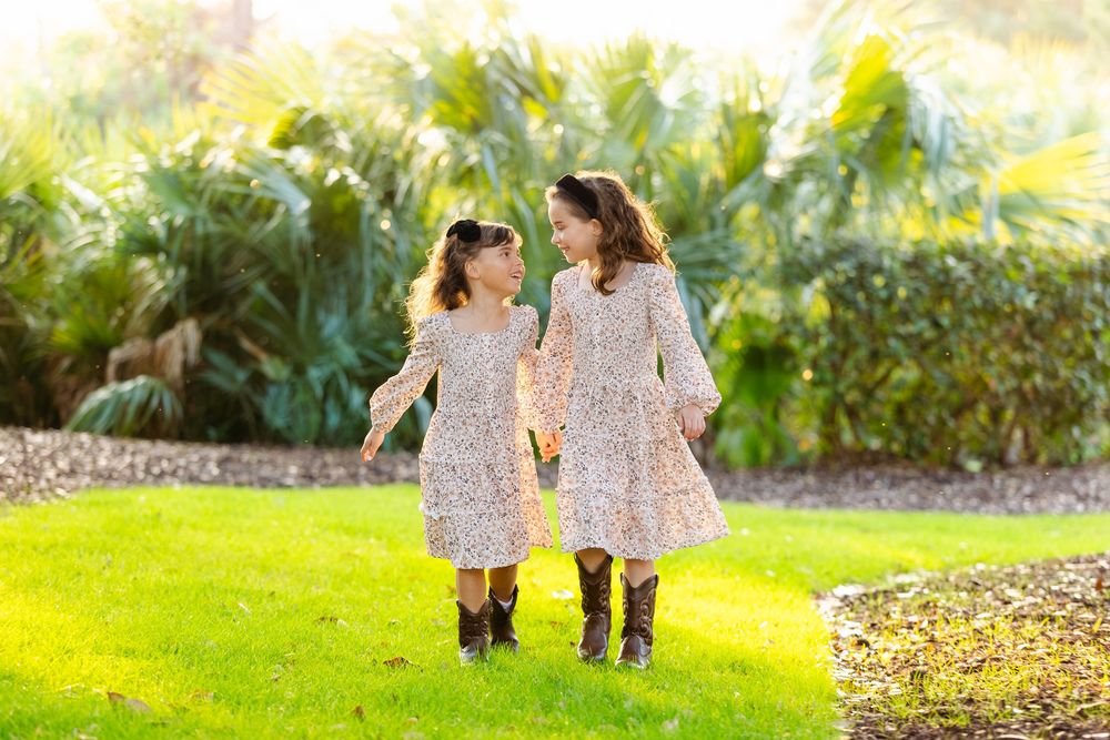 Two young sisters in matching floral dresses and cowboy boots walking together on a sunny green lawn.