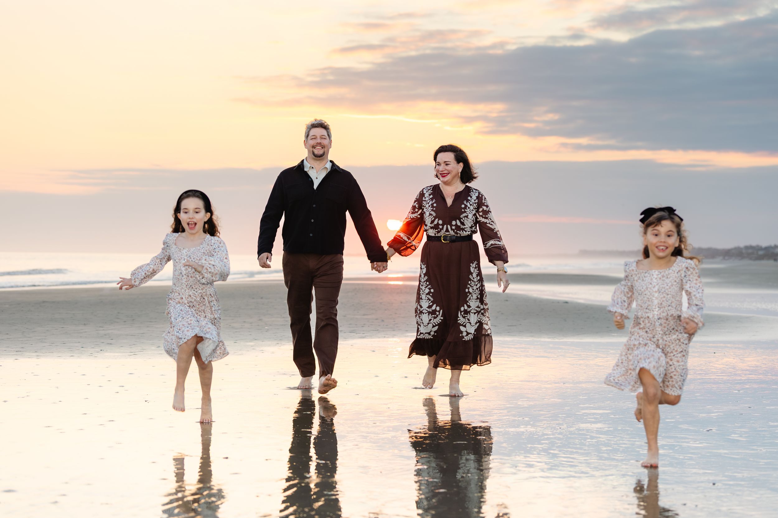 Family of four holding hands and running barefoot on a beach at sunset with reflective wet sand.