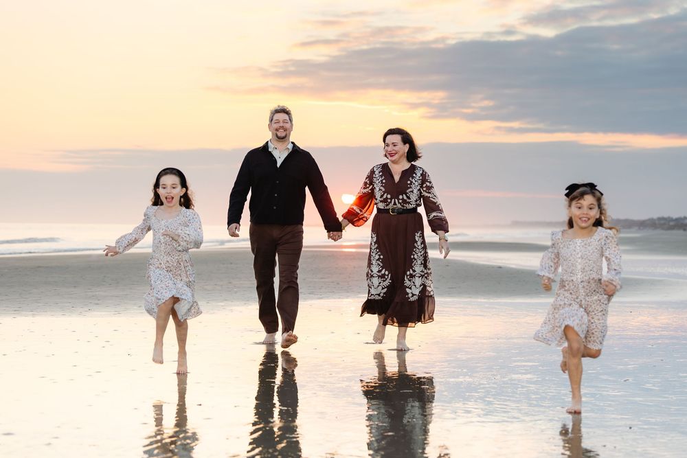 Family of four holding hands and running barefoot on a beach at sunset with reflective wet sand.