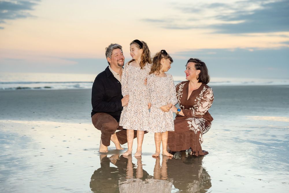 Happy family of four posing together on a beach at sunset, smiling and embracing near the shoreline.