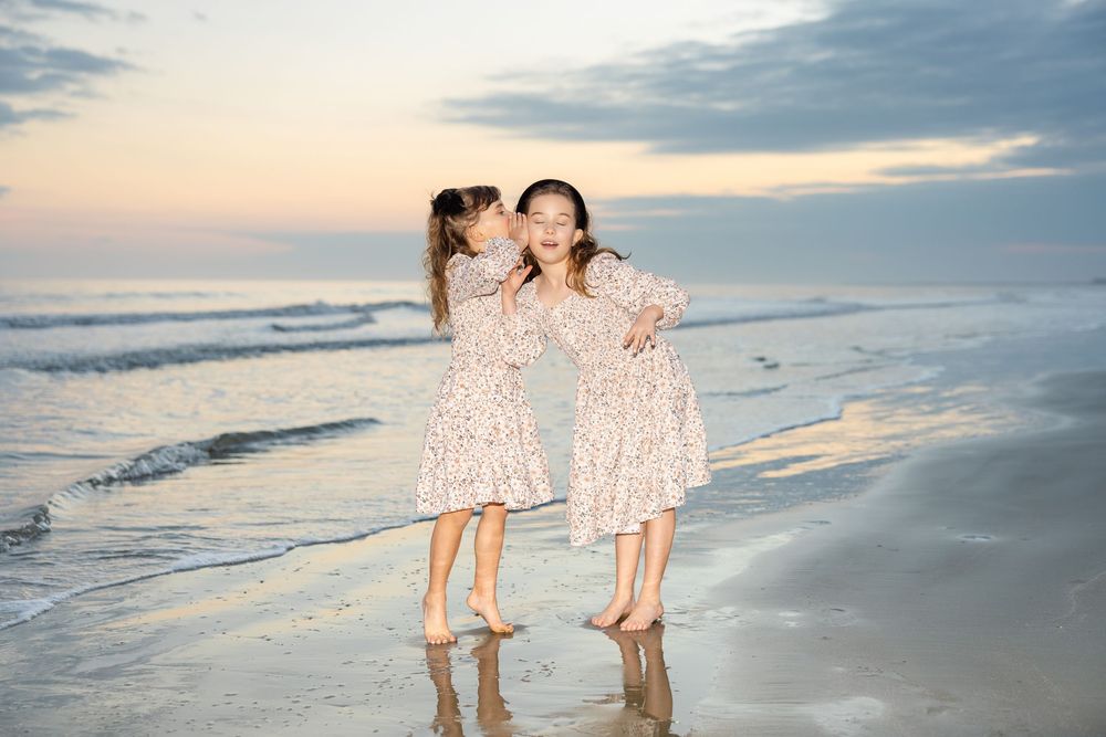 Two girls in matching floral dresses playing on a beach at sunset, one whispering to the other.