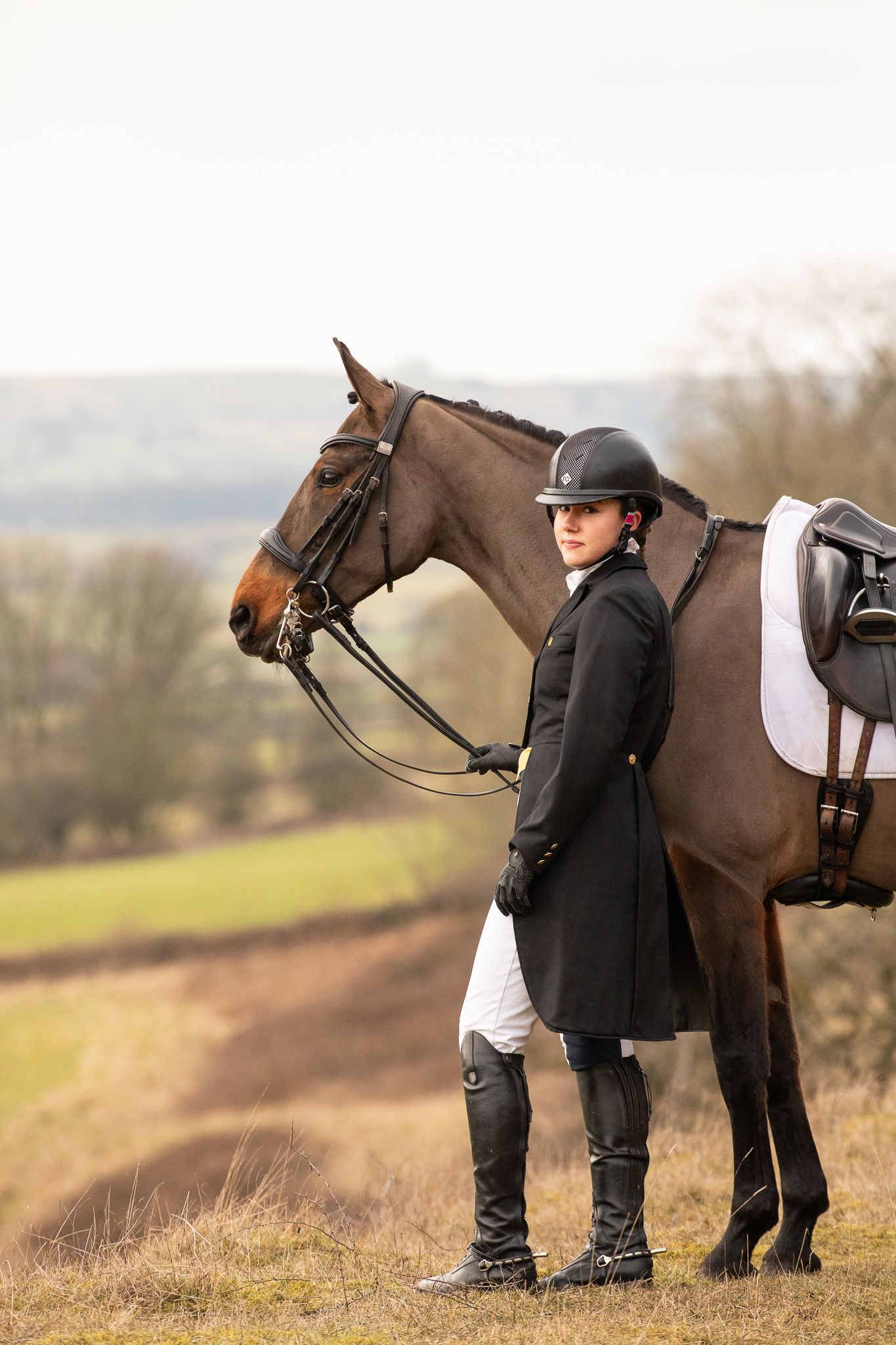 An equestrian in traditional riding attire stands with a saddled brown horse on a hillside overlooking rural countryside.