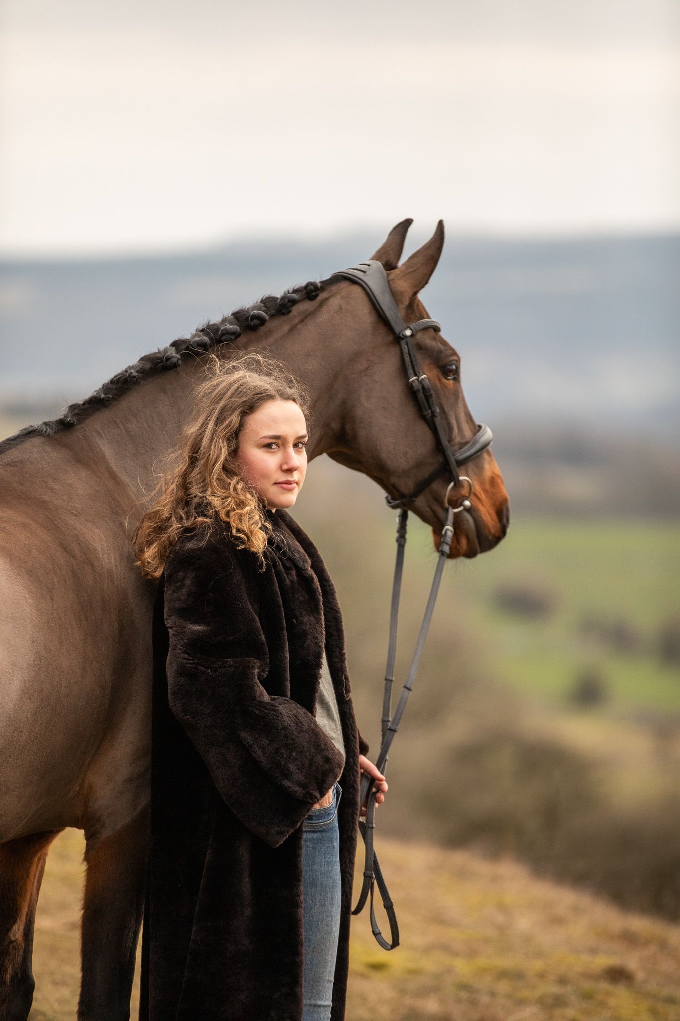 A beautiful white horse stands with its handler in brown winter attire on a hillside with dried vegetation.