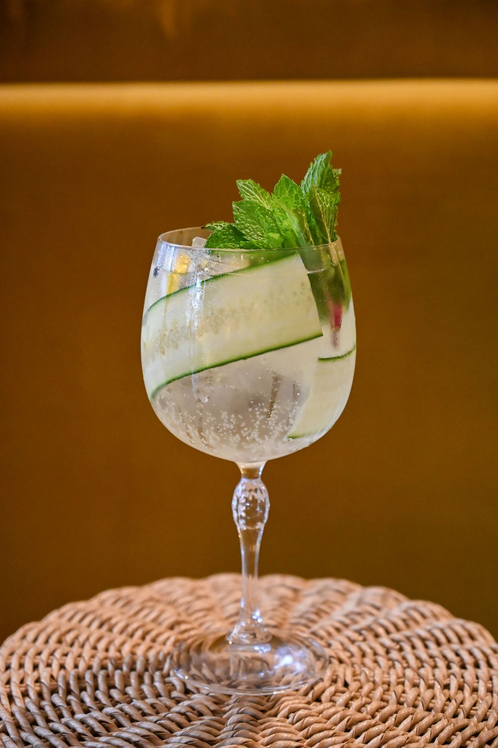 A refreshing gin and tonic cocktail garnished with fresh mint leaves and cucumber on a woven rattan placemat.