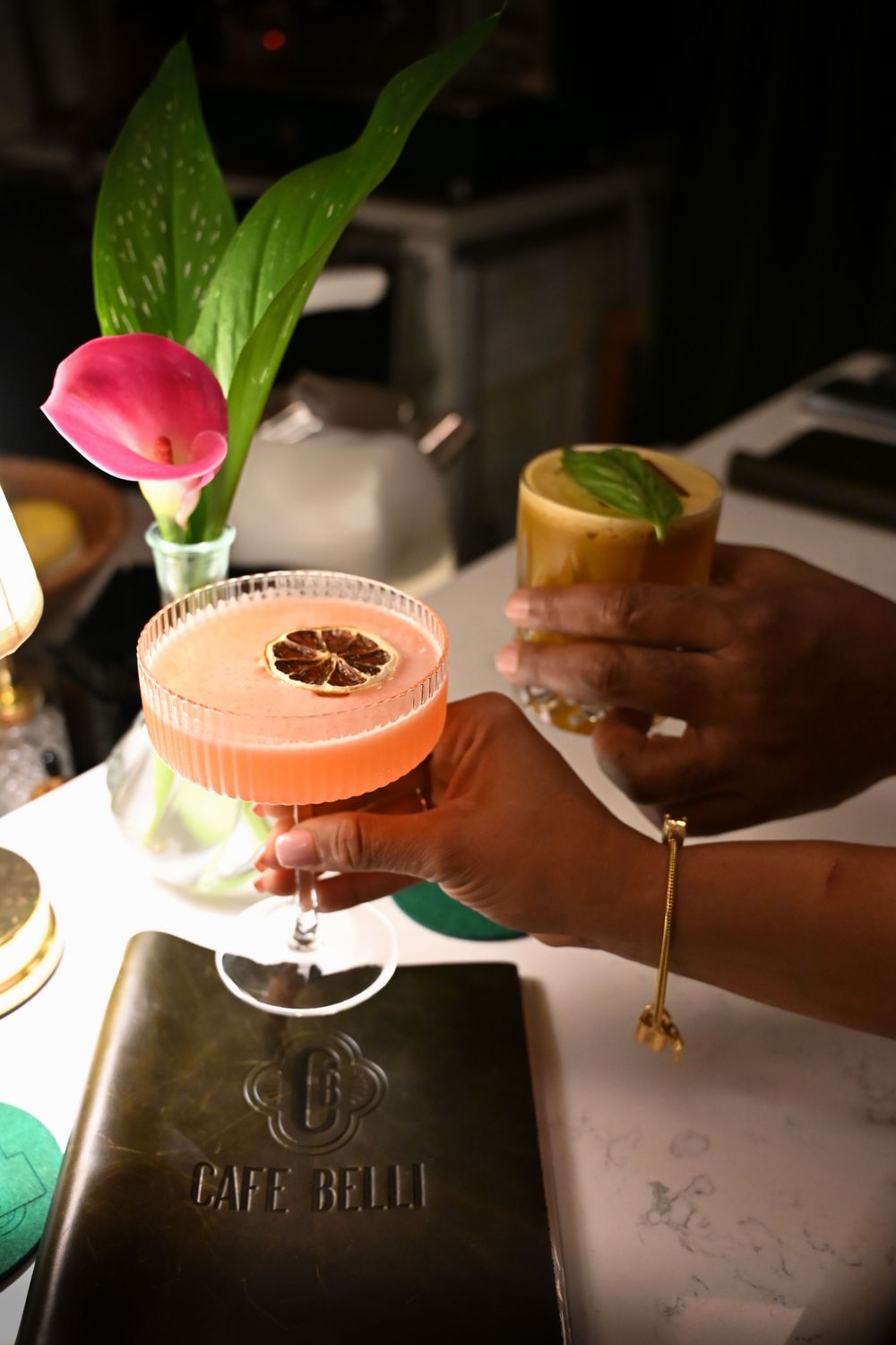 Three pink cocktails in martini glasses garnished with fresh leaves are served on a dark table in dim lighting.