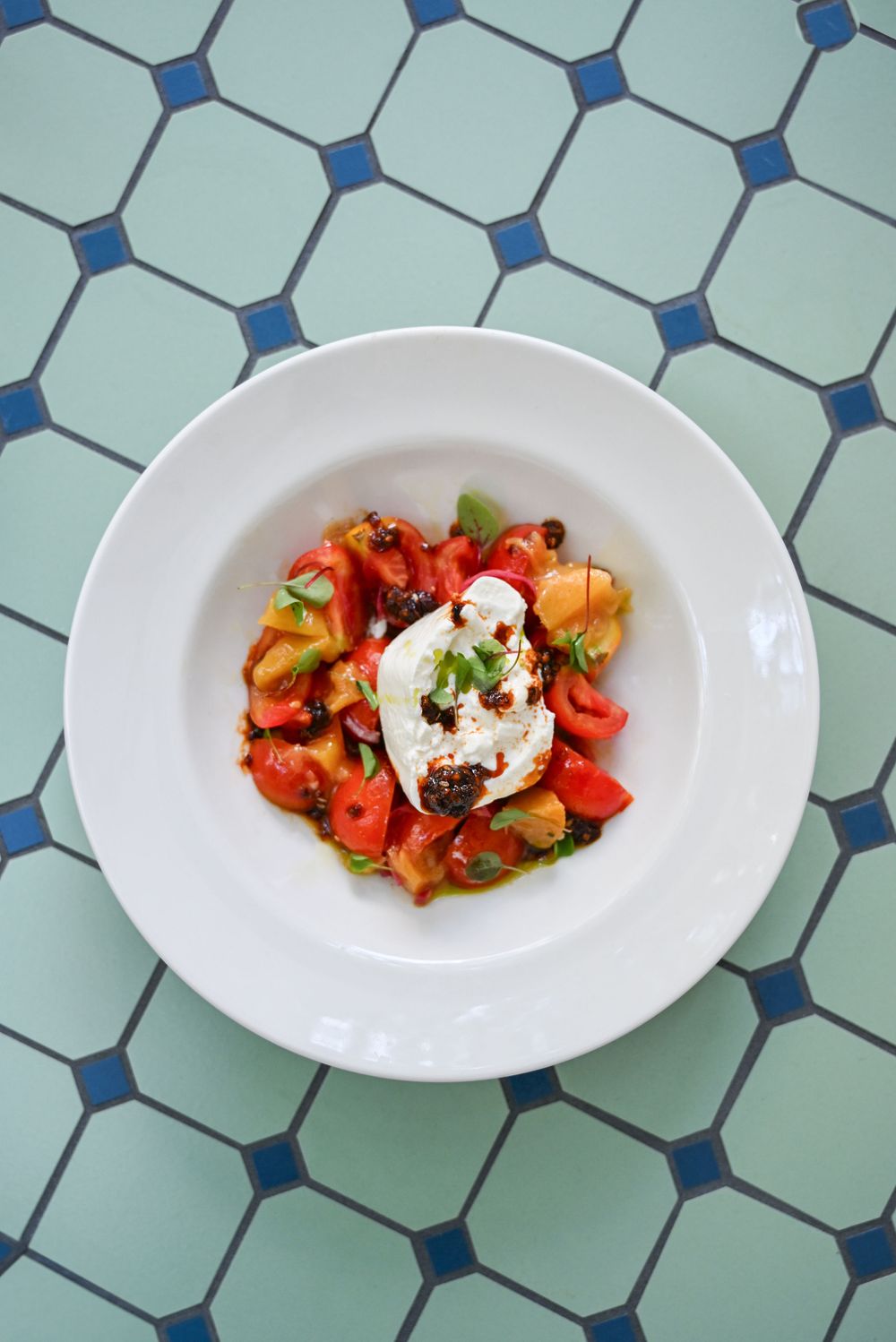 Three white plates of fresh burrata salad with tomatoes and herbs arranged on a blue geometric tiled surface.