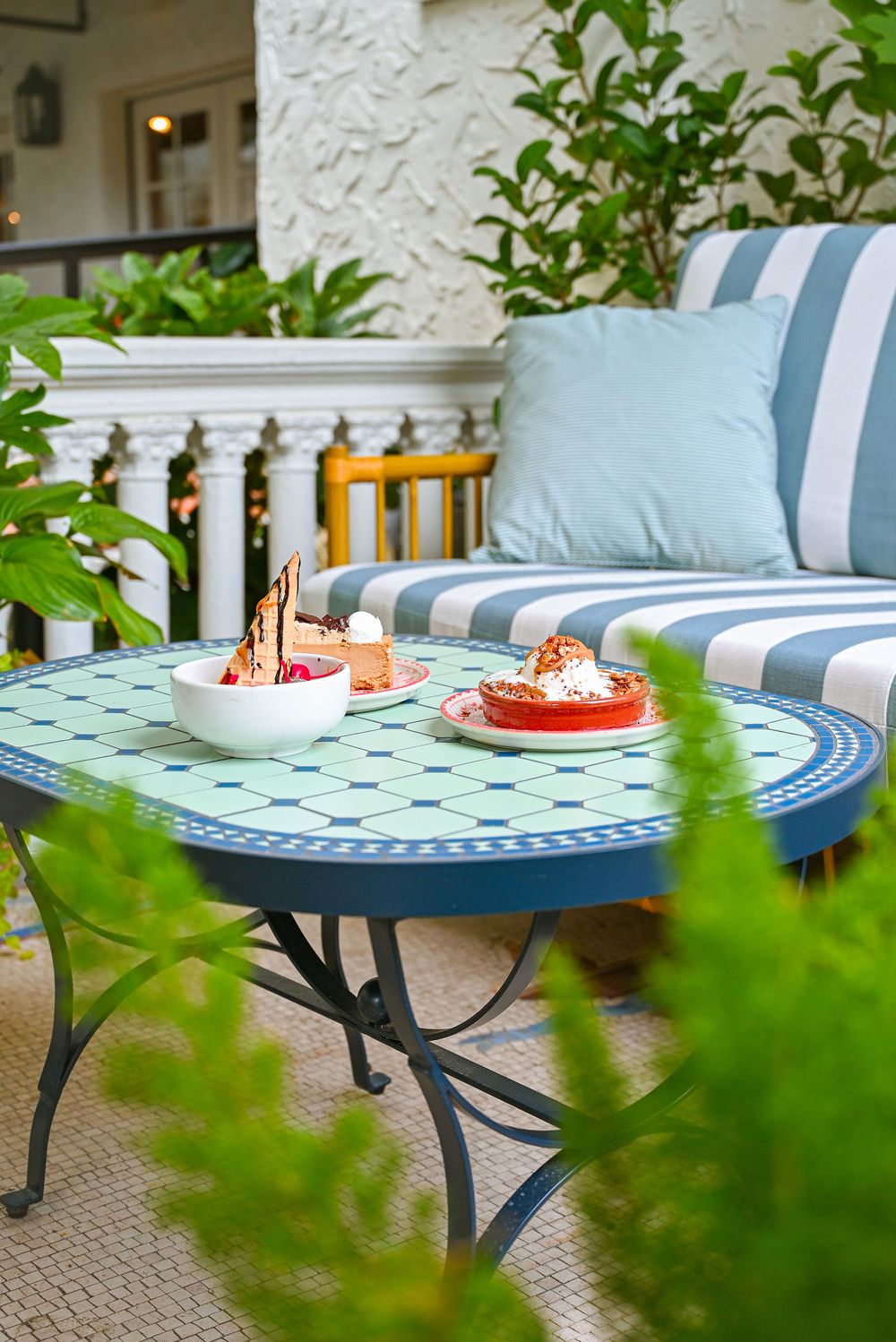 A blue patterned outdoor table with striped cushions on patio furniture is framed by green foliage in the foreground.
