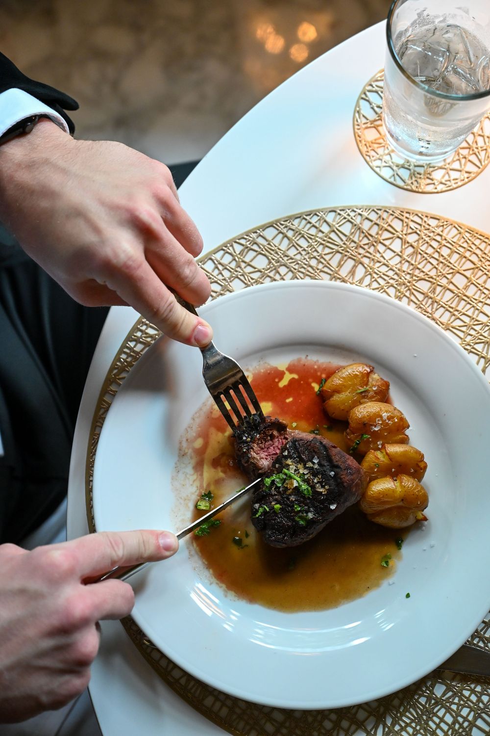 Hands cutting into a seared steak with roasted potatoes on an elegant white plate with gold accents and warm lighting.