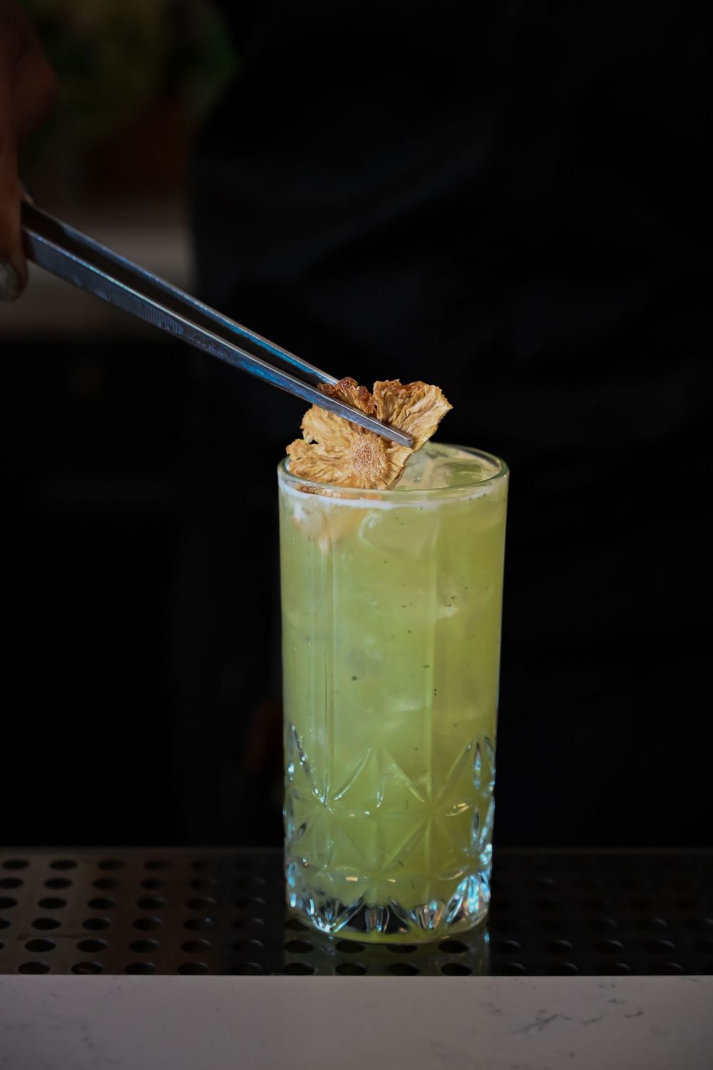 A tall glass of green cocktail garnished with a morsel held by chopsticks against a dark background.