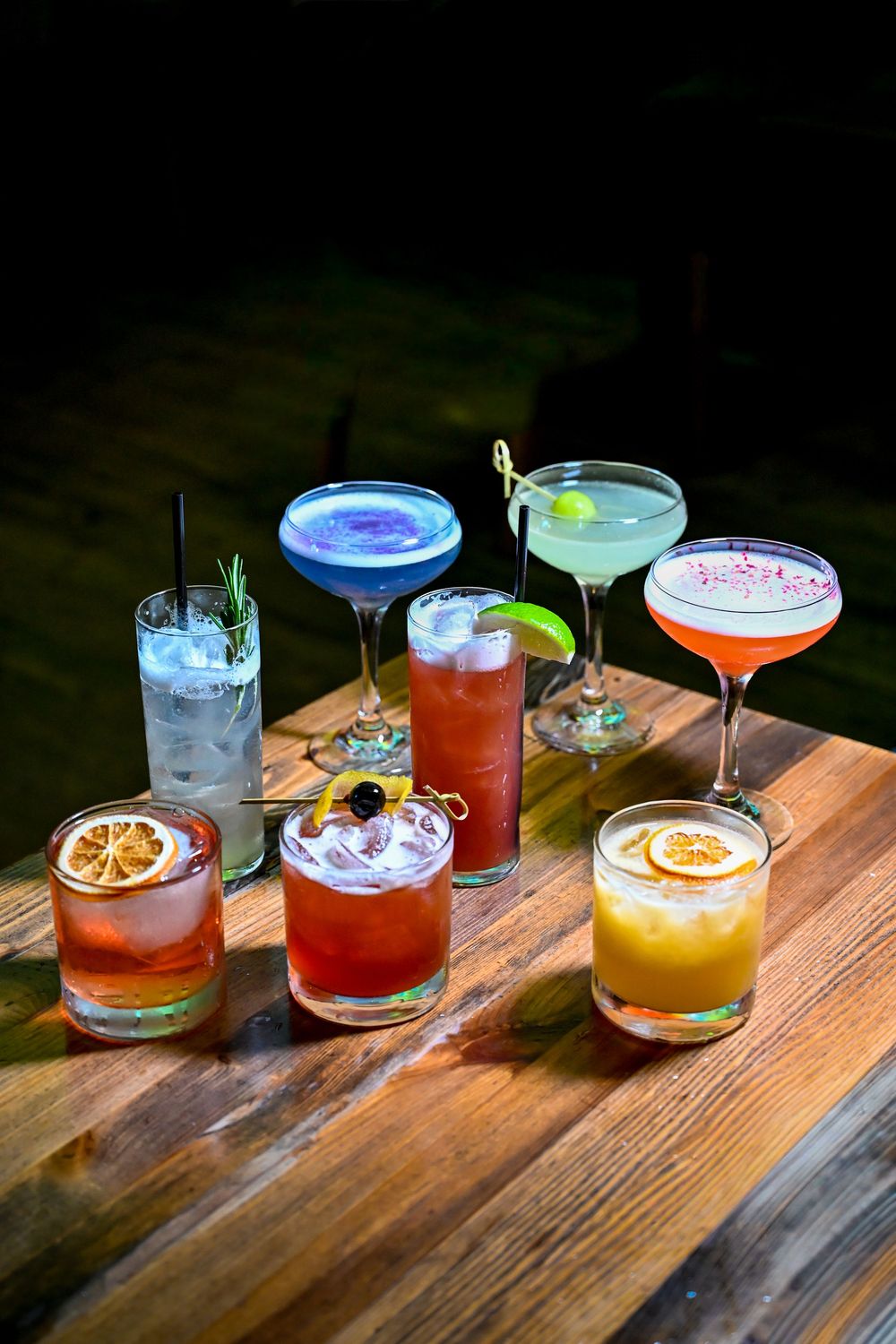 Seven colorful cocktails displayed on a wooden table, featuring garnishes like citrus slices, herbs, and berries.