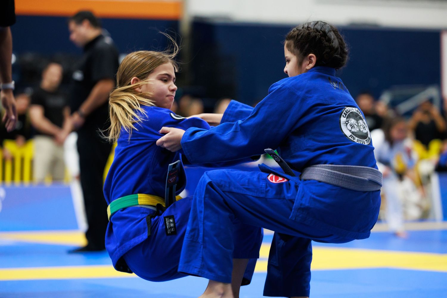 Martial artists in blue gis demonstrate grappling techniques during a competition.