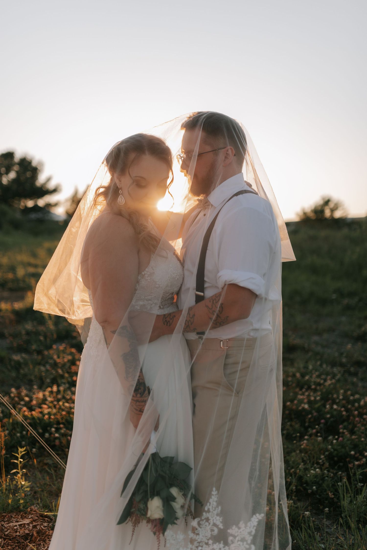 Bride and groom under veil at golden hour sunset in lush green field.