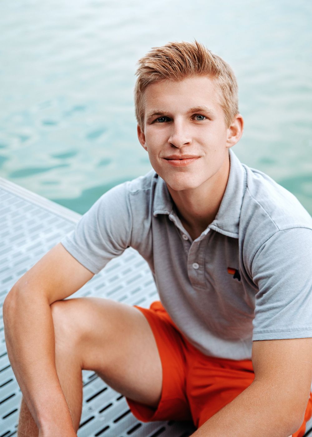 Young person in a grey polo shirt and red shorts sitting on a dock by turquoise water on a sunny day.