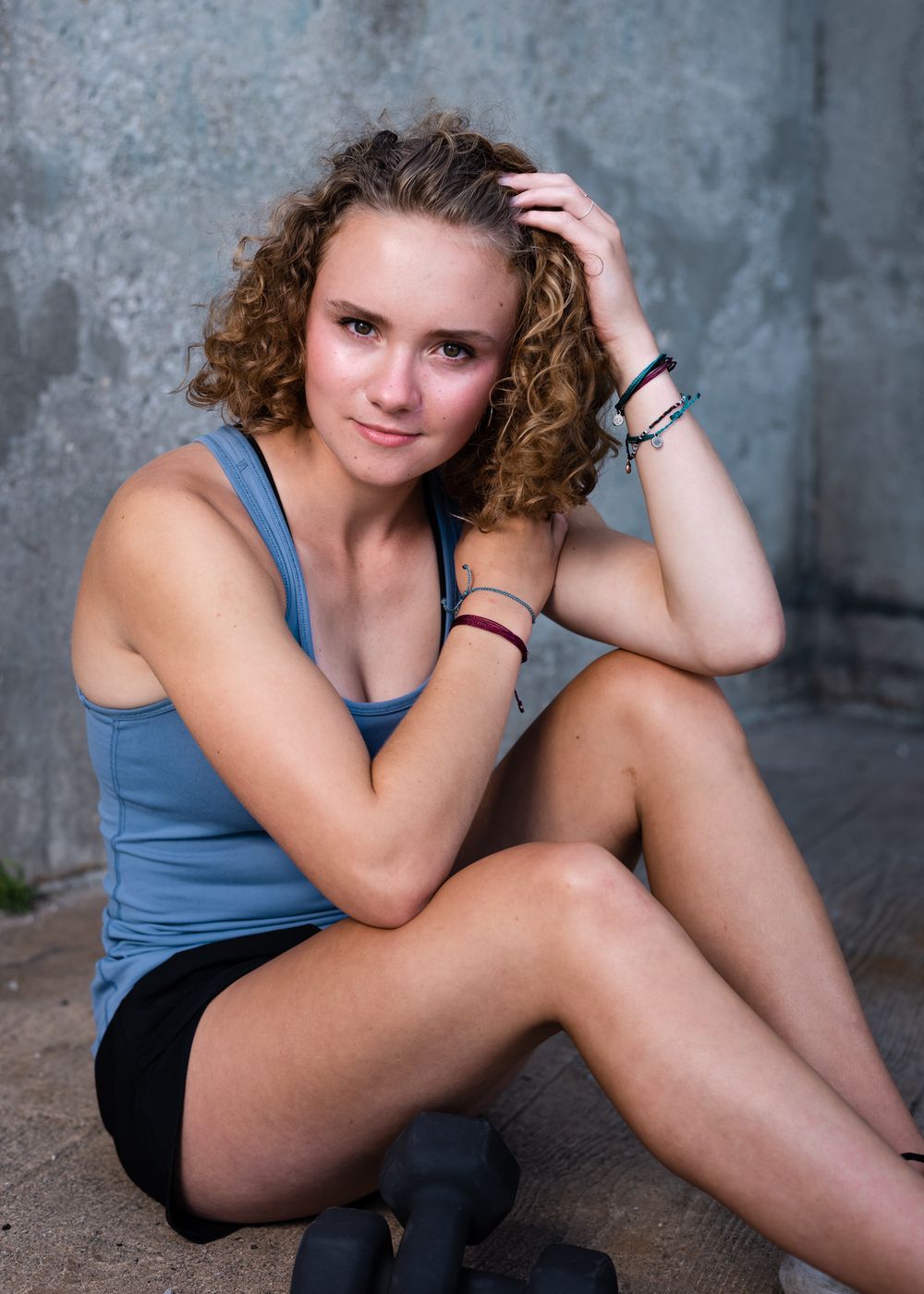 Athletic person in a blue tank top and black shorts poses while sitting against a textured concrete wall background.