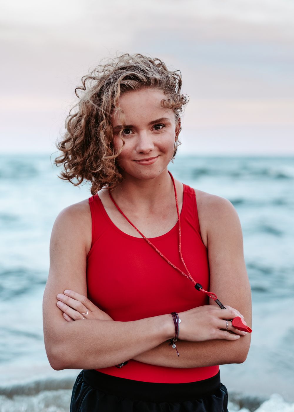 A person with curly hair wearing a red tank top poses with crossed arms at the beach during sunset with ocean waves behind.