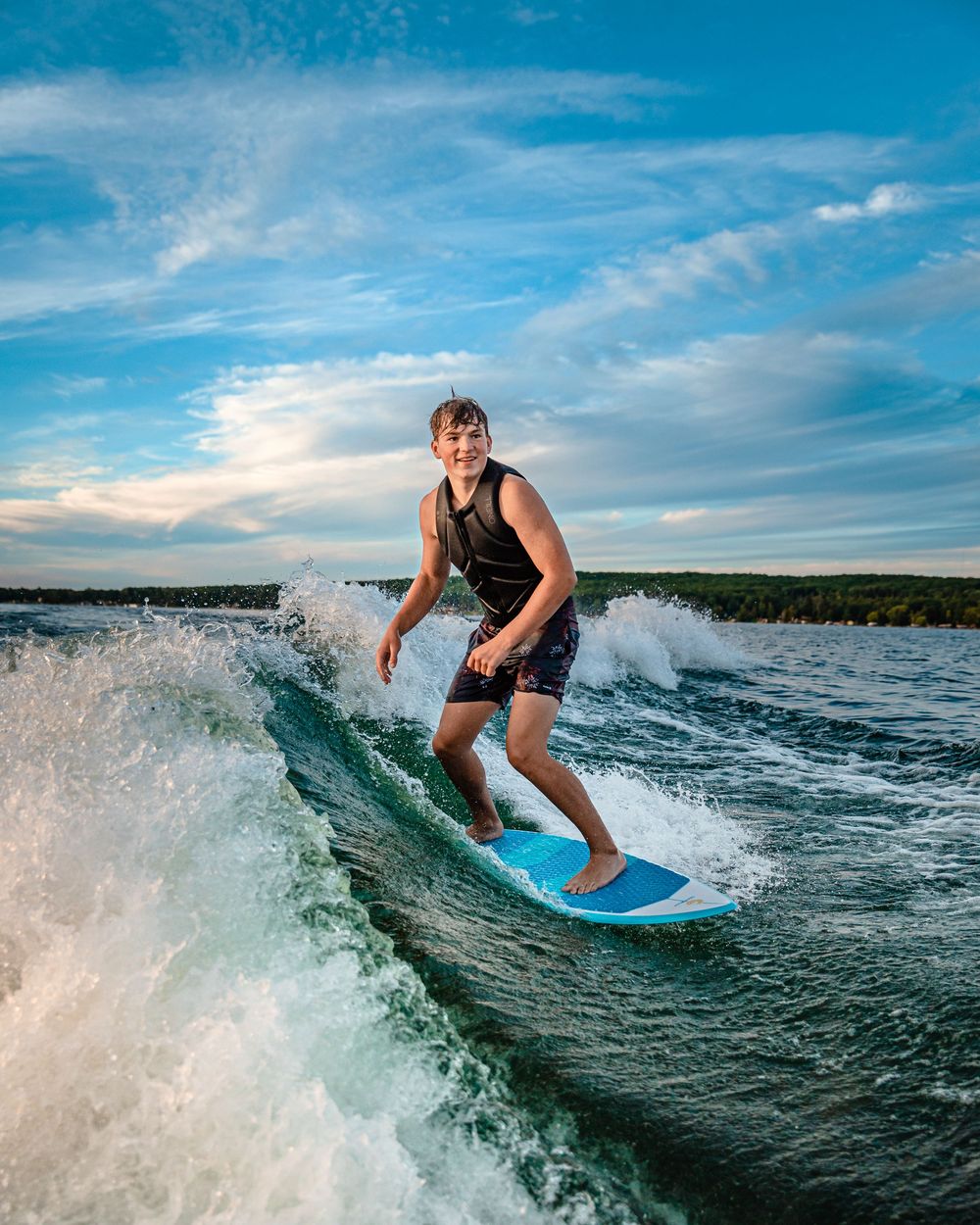 Wakeboarder riding a blue board through splashing waves on a lake during sunset with dramatic clouds in the sky.