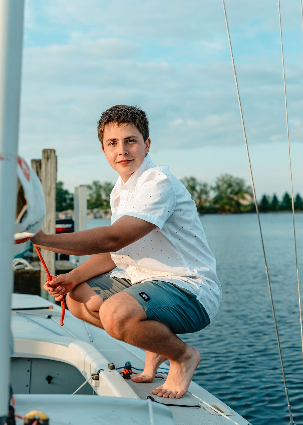 A young person in a white shirt sits barefoot on the edge of a sailboat on a calm lake during a sunny day.