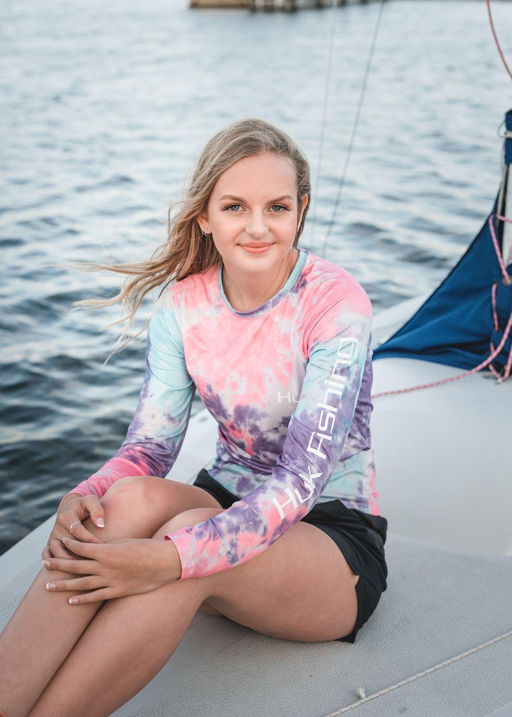 Someone relaxing on a sailboat wearing a pink and purple tie-dye long sleeve shirt and black shorts on the water.