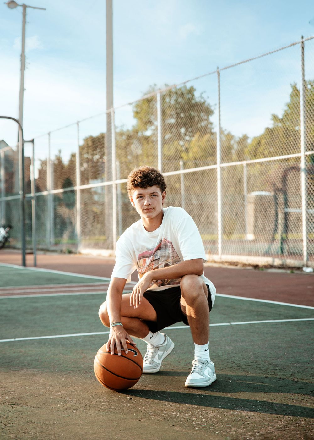 Basketball player crouches on outdoor court holding ball while wearing white t-shirt and shorts in sunny setting.