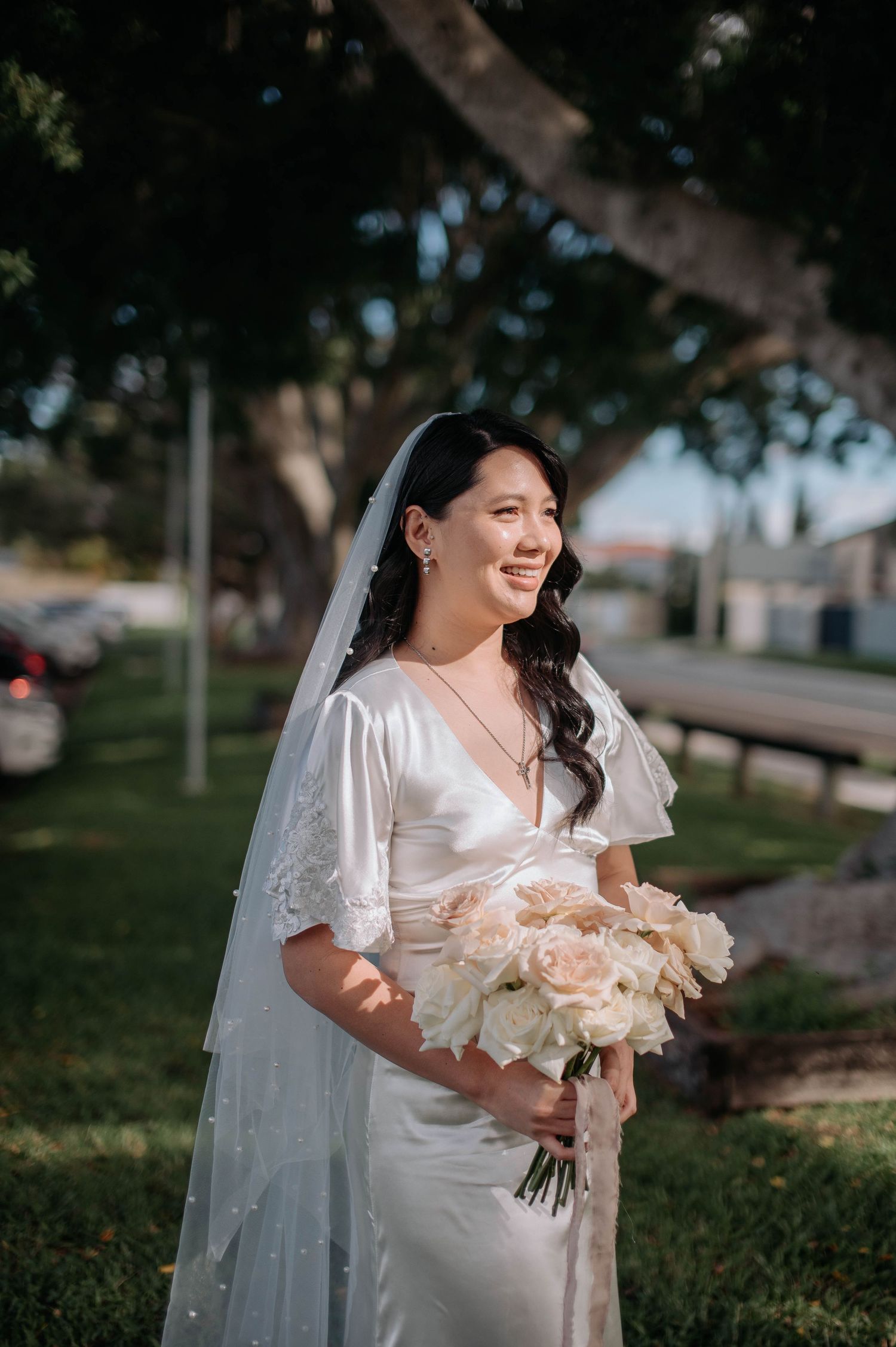 A radiant bride in a flowing white dress and veil holds a pale pink bouquet while standing outdoors on a sunny day.
