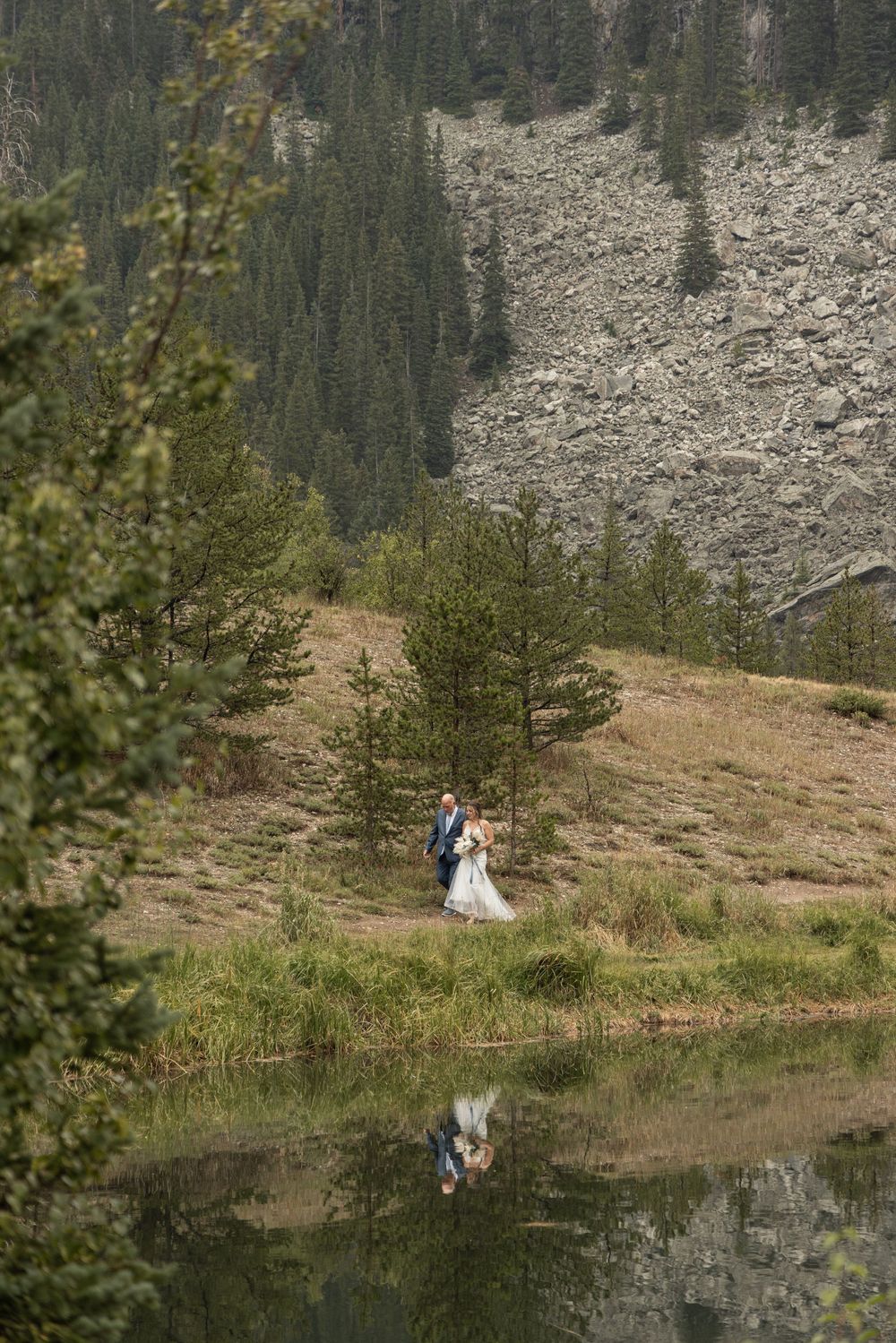A scenic mountain landscape with evergreen trees and a small lake reflecting two figures standing on a grassy slope.