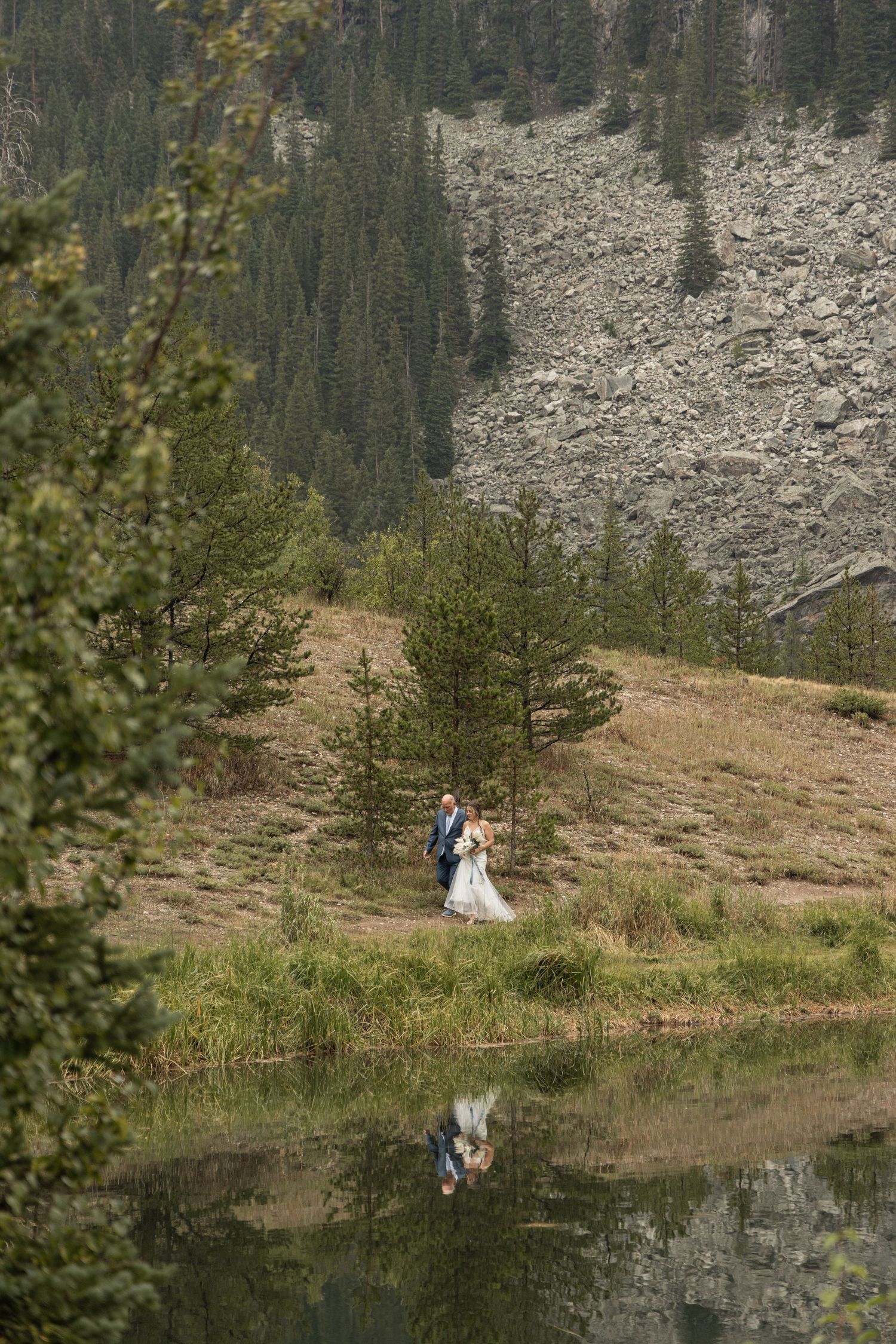 A scenic mountain landscape with evergreen trees and a small lake reflecting two figures standing on a grassy slope.