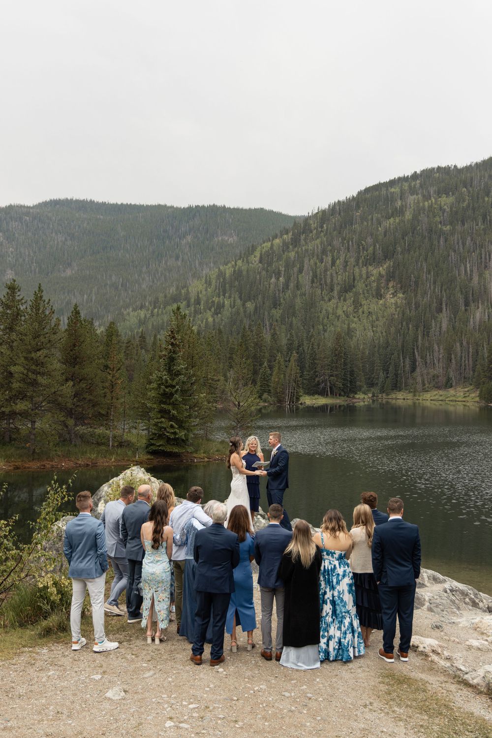 Outdoor mountain lake wedding ceremony with guests gathered on rocky shore surrounded by evergreen forest and hills.