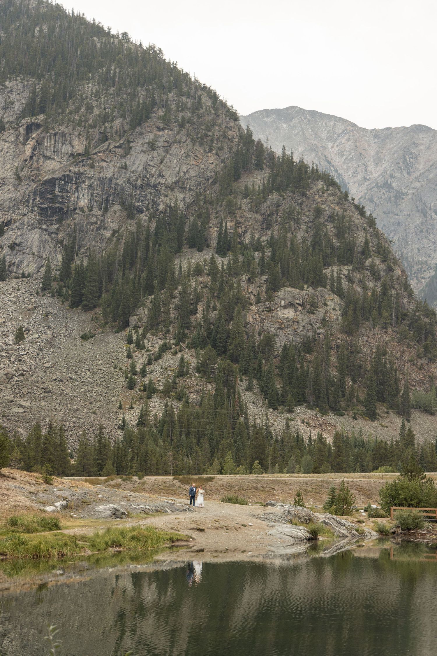 A narrow winding mountain path leads to a serene alpine lake surrounded by steep rocky cliffs and evergreen forest.