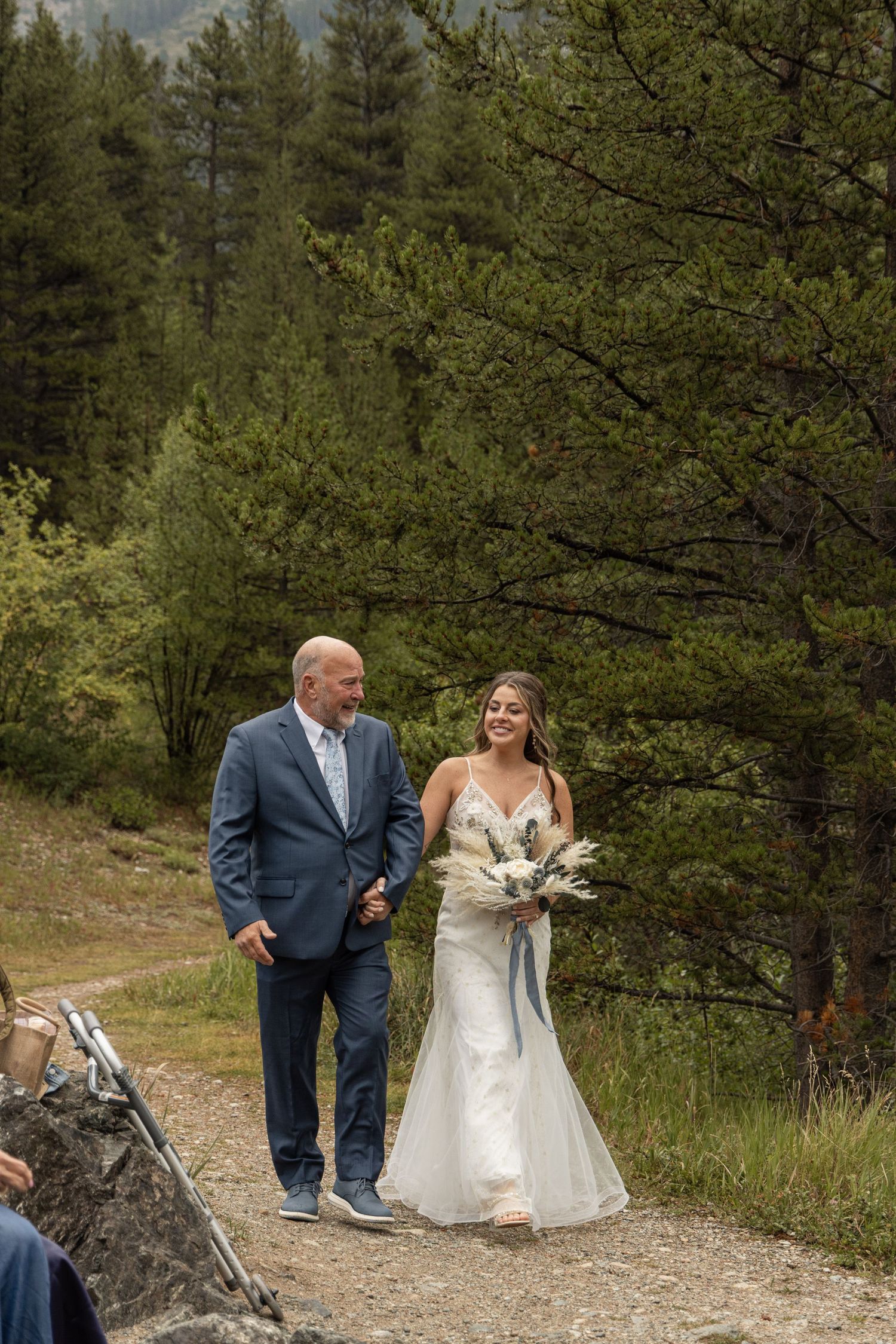 A couple shares a joyful moment on a scenic mountain path surrounded by pine trees and natural greenery.