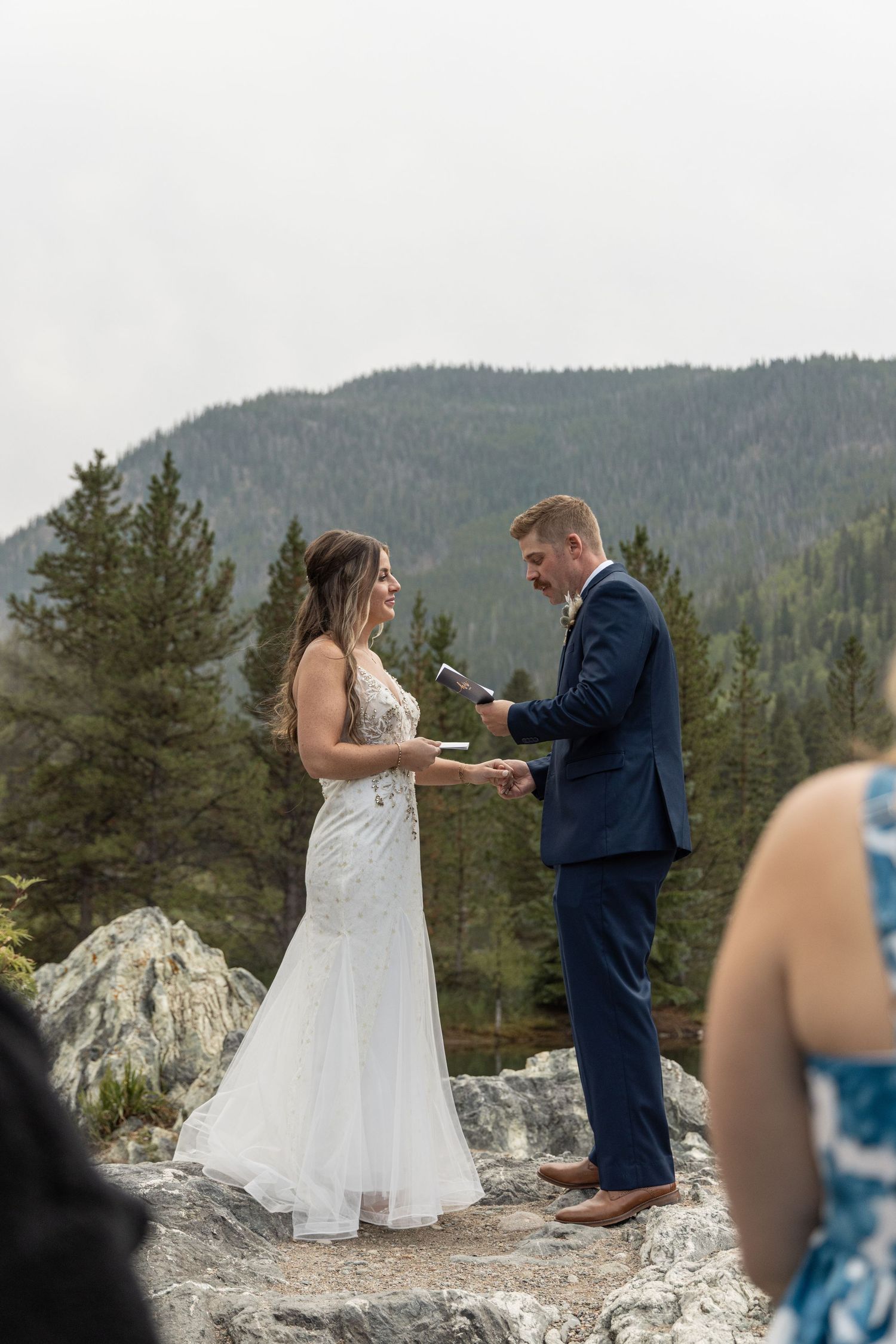 A couple in wedding attire shares a tender moment on a rocky mountain overlook with evergreen trees in the background.