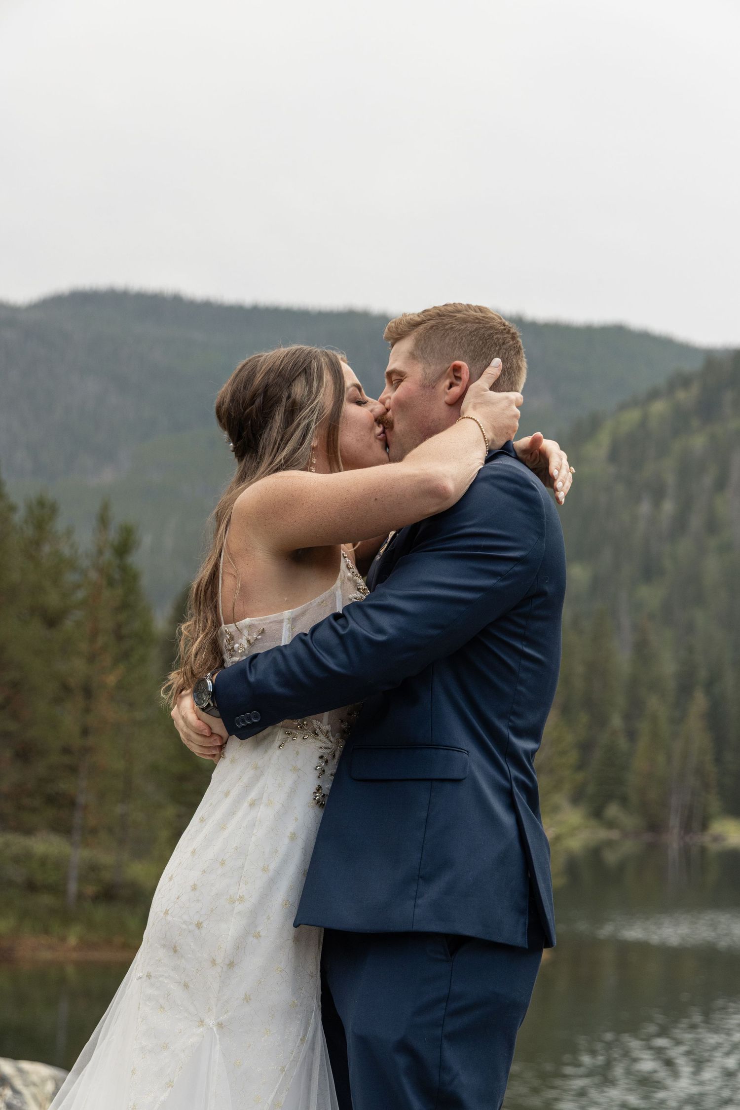 Newlywed couple shares romantic kisses by a serene mountain lake with evergreen trees in the background.