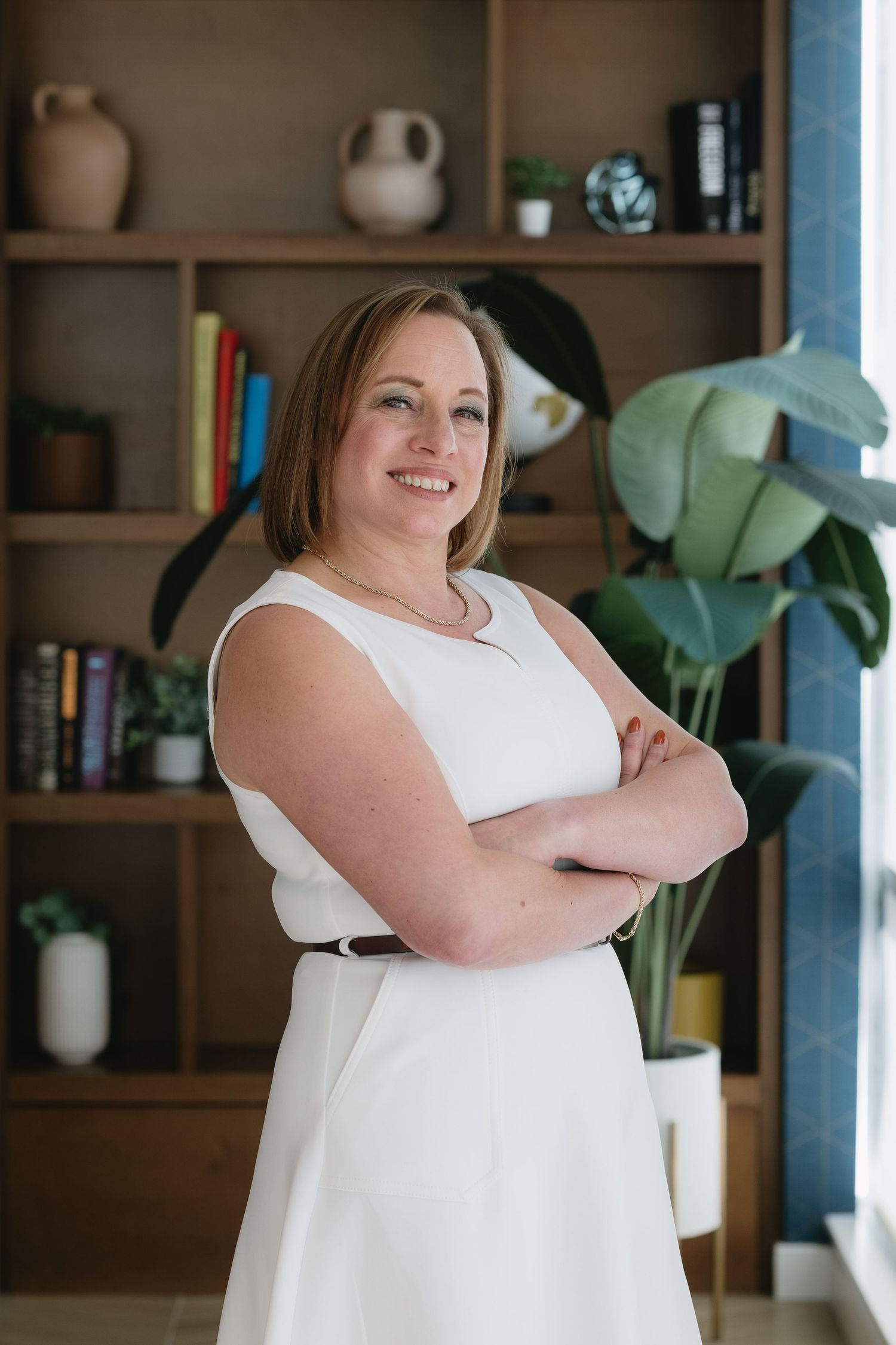 Interior portrait session featuring someone in white dress against shelved wall with plants.