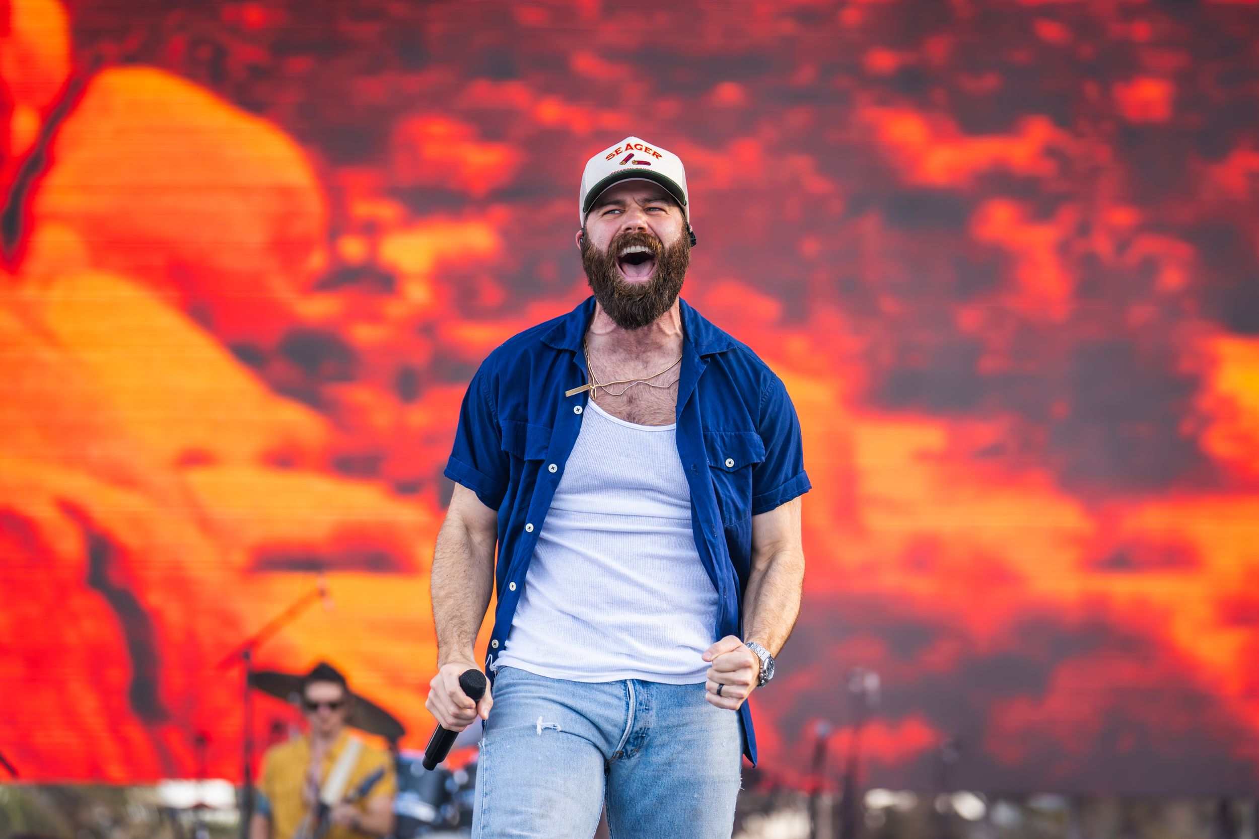 Concert performer in white tank top and blue shirt against vibrant orange backdrop.