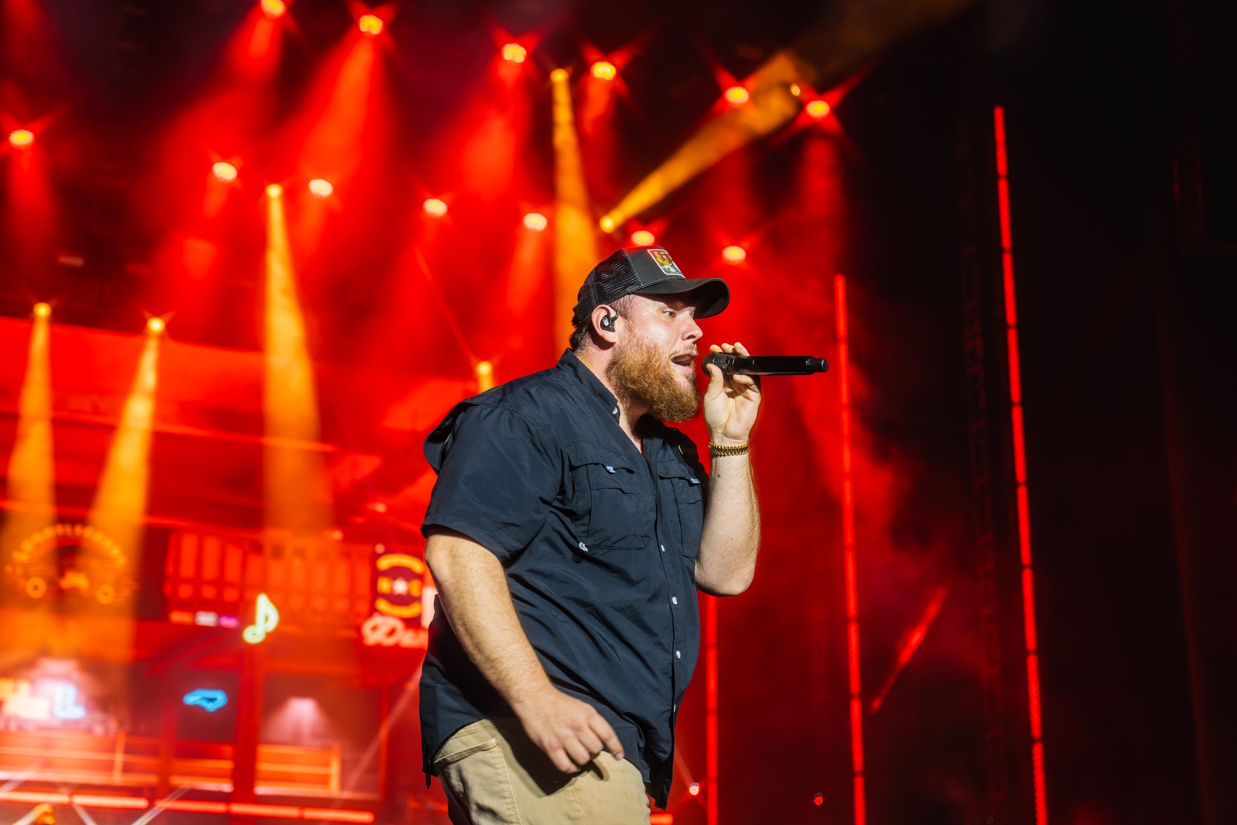 Stage performer in dark clothing singing into microphone against red concert lighting.