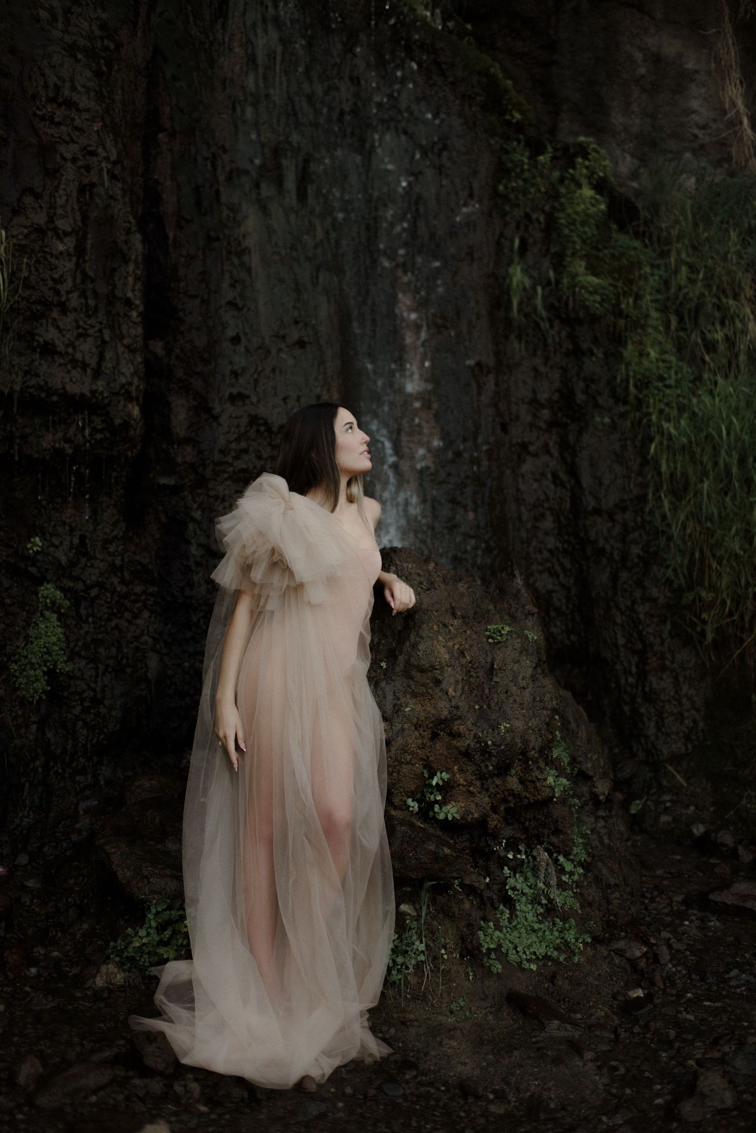 An ethereal figure in a sheer cream gown poses dramatically against a dark rocky cliff face.