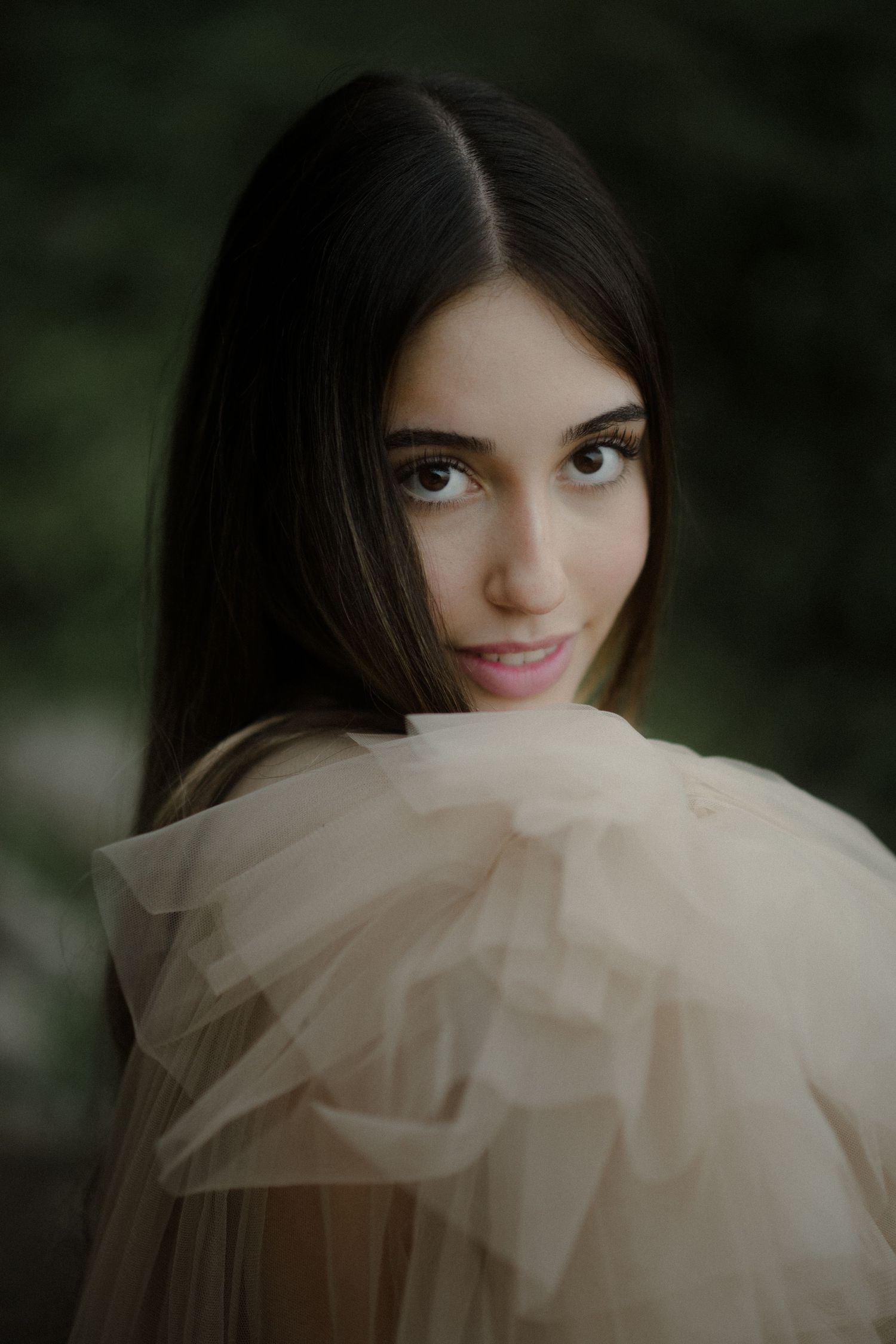 Woman in soft tulle fabric poses against dark background with romantic lighting.