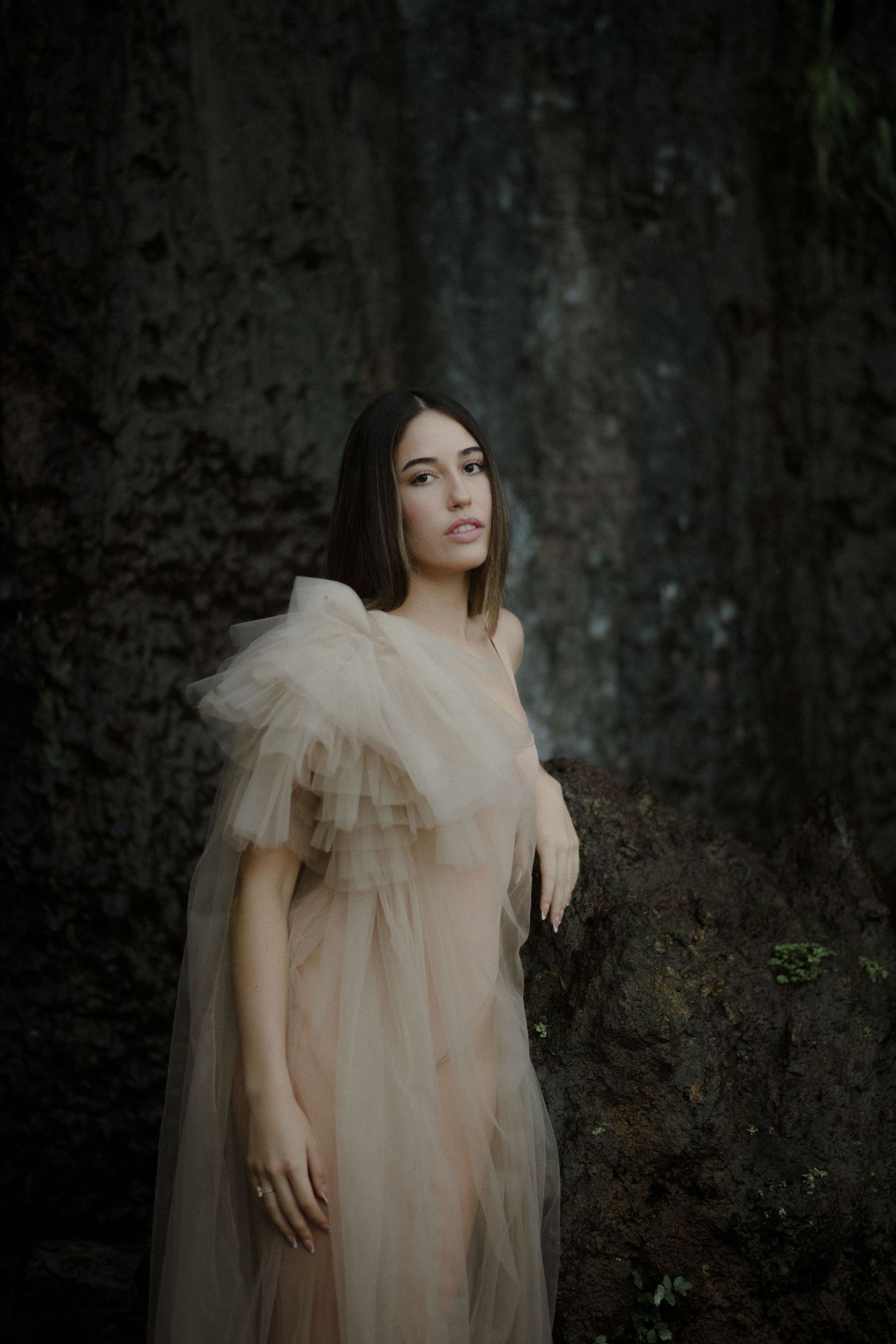 A moody portrait against a dark stone wall wearing a romantic tulle dress in pale cream.