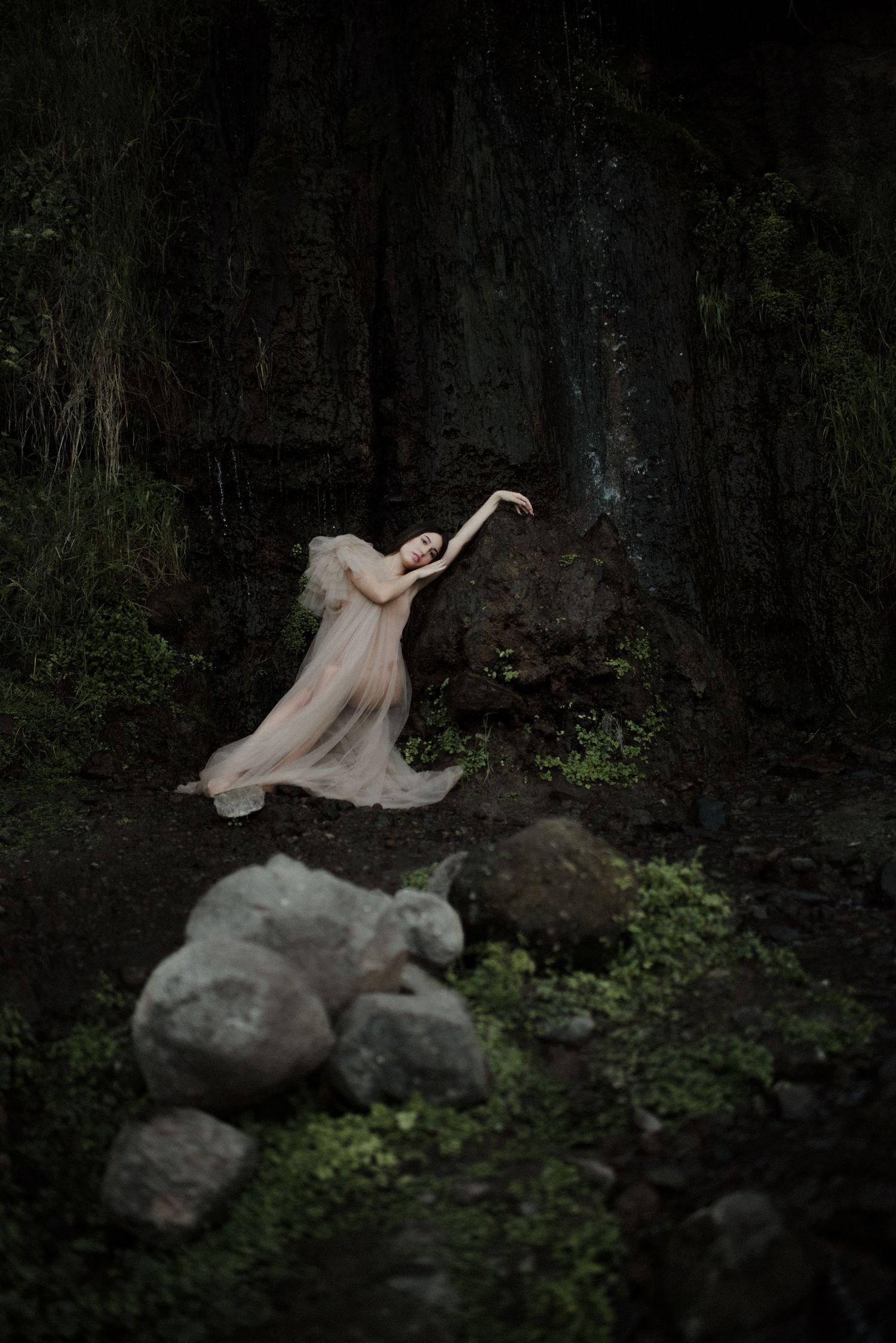 Ethereal figure in flowing dress lying among dark forest foliage and stone.