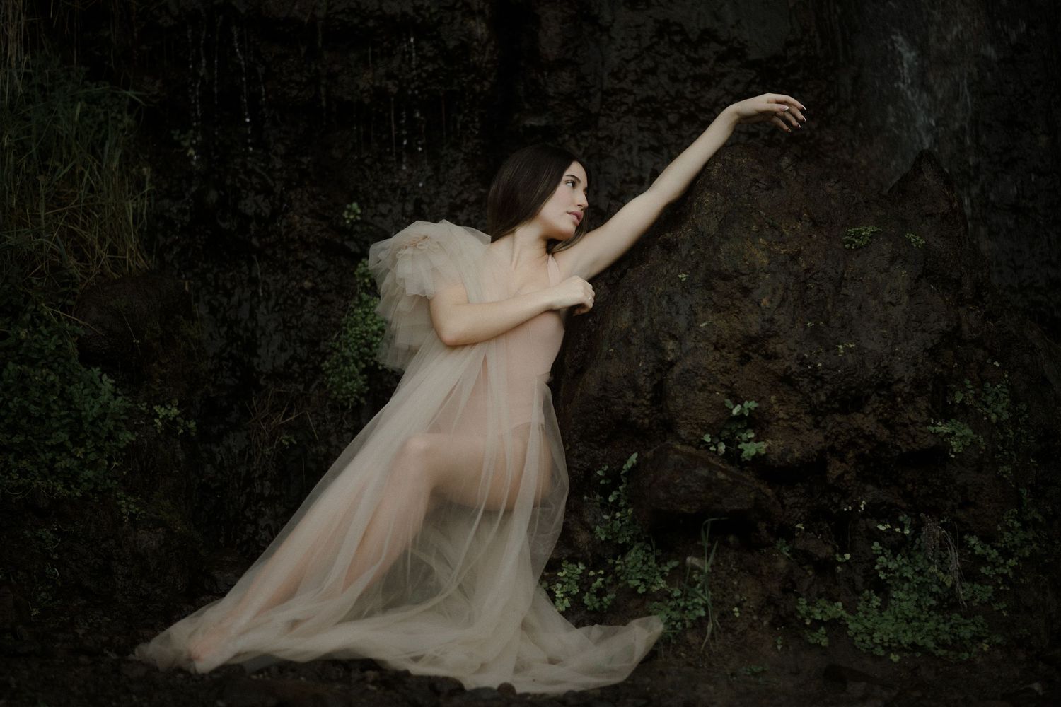 A graceful dance-like pose in a sheer cream dress against a dramatic dark rocky backdrop.