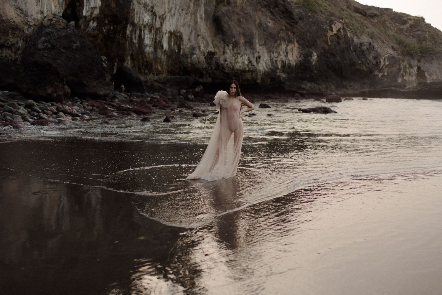 Figure in flowing dress walks along rocky beach shoreline at sunset.