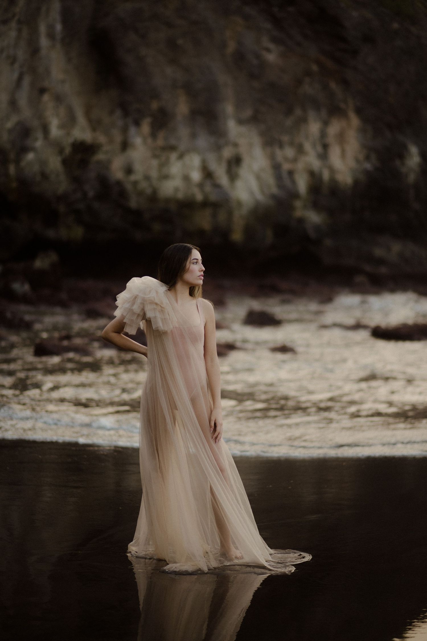 Ethereal figure in flowing blush dress stands gracefully near dark cliffside at dusk.