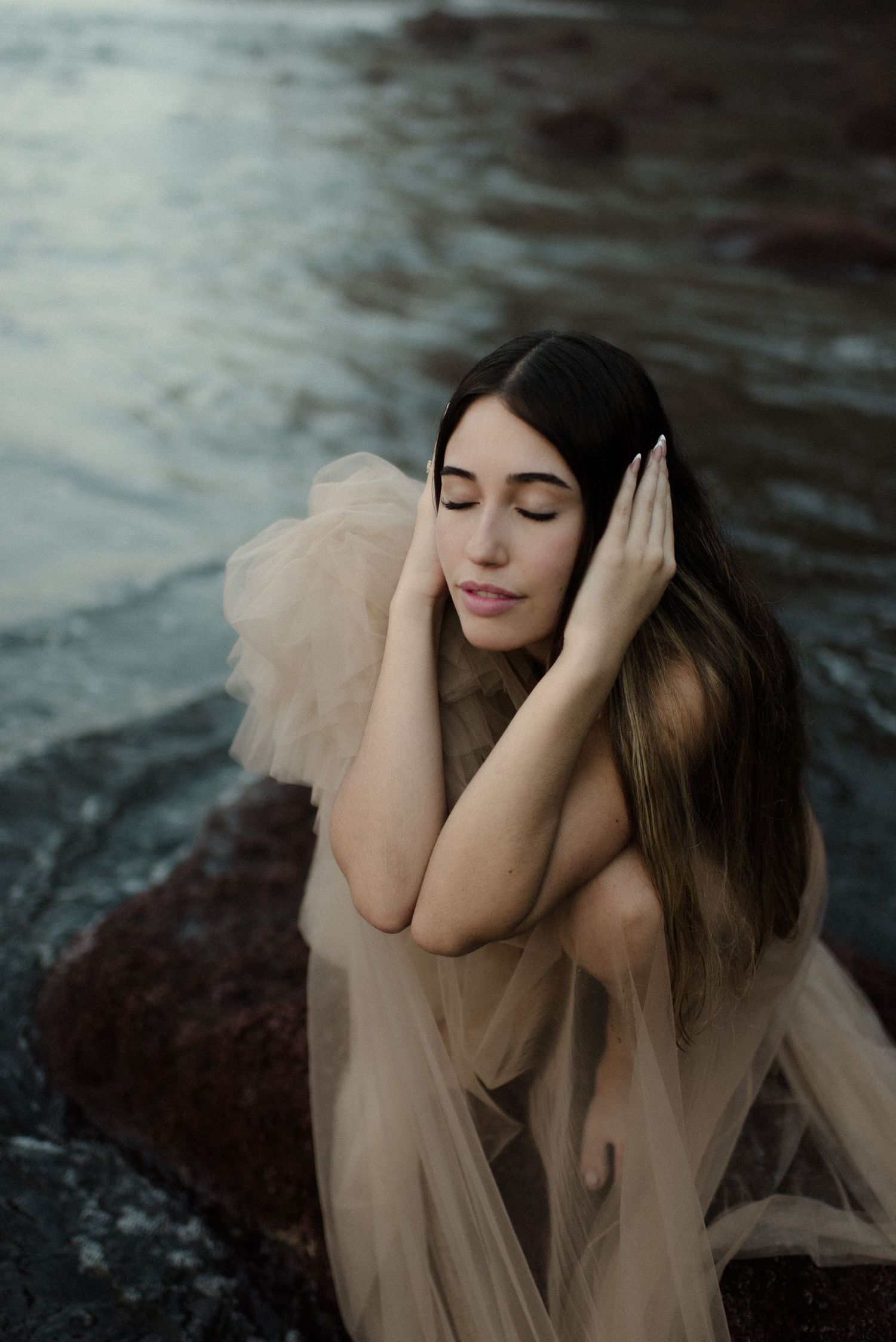 Woman in beige tulle dress poses gracefully on rocky beach landscape.