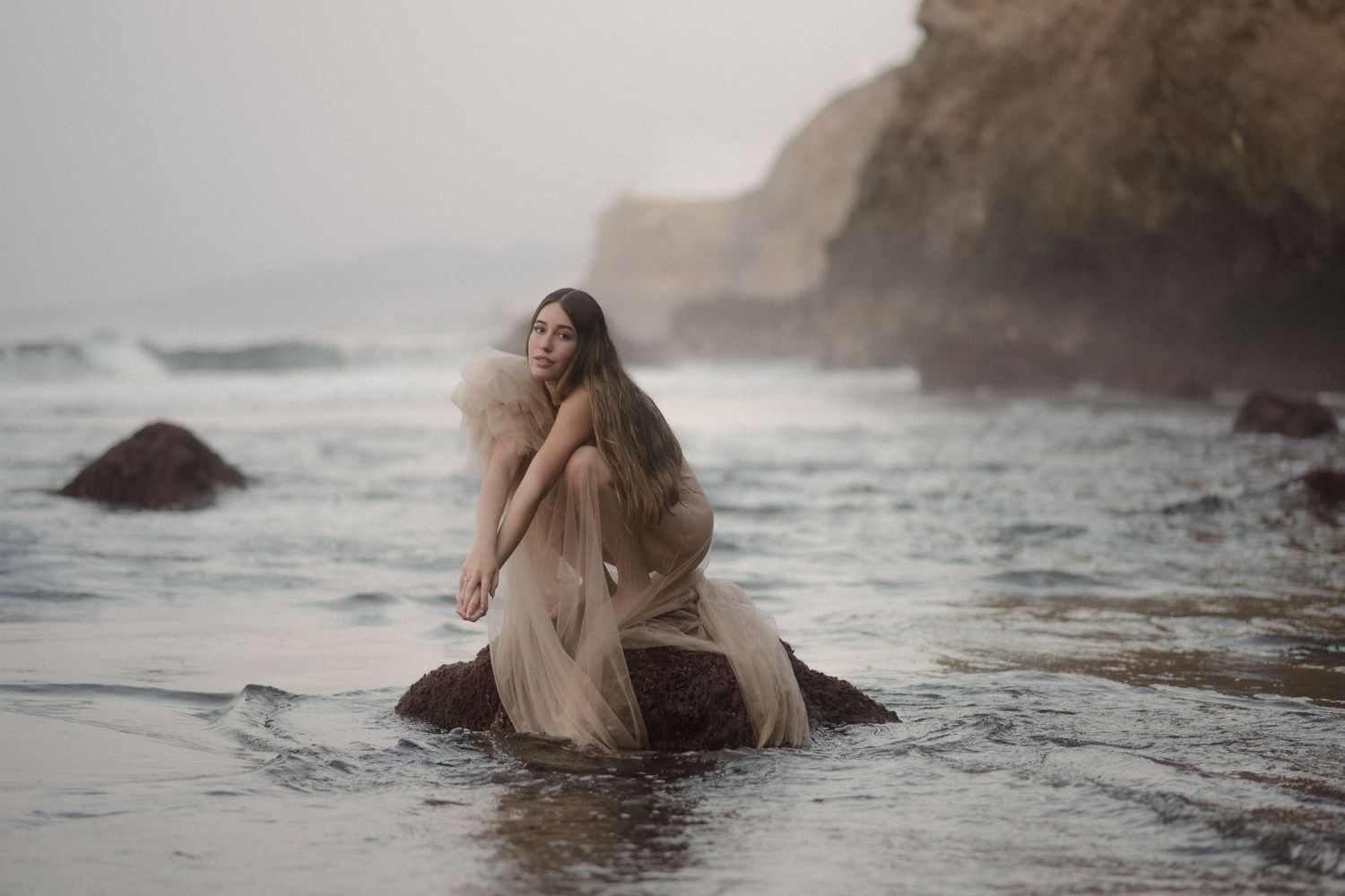 Ethereal figure in flowy dress poses on coastal rocks during misty sunset at rugged beach.