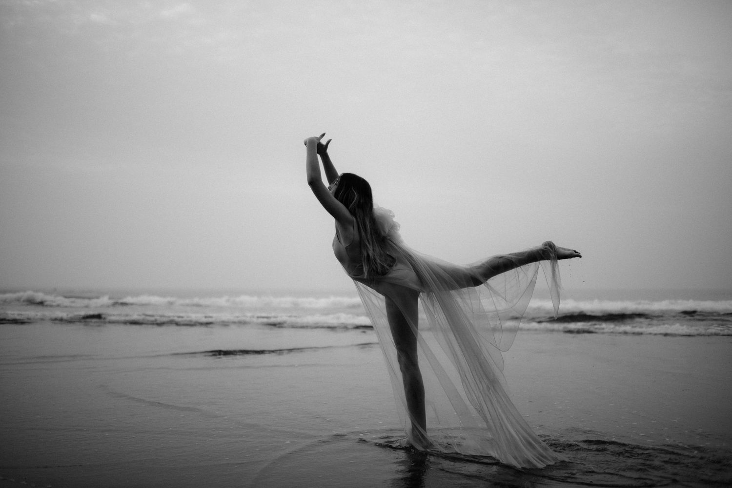 Ballet dancer practicing elegant poses along the shoreline in black and white photography.