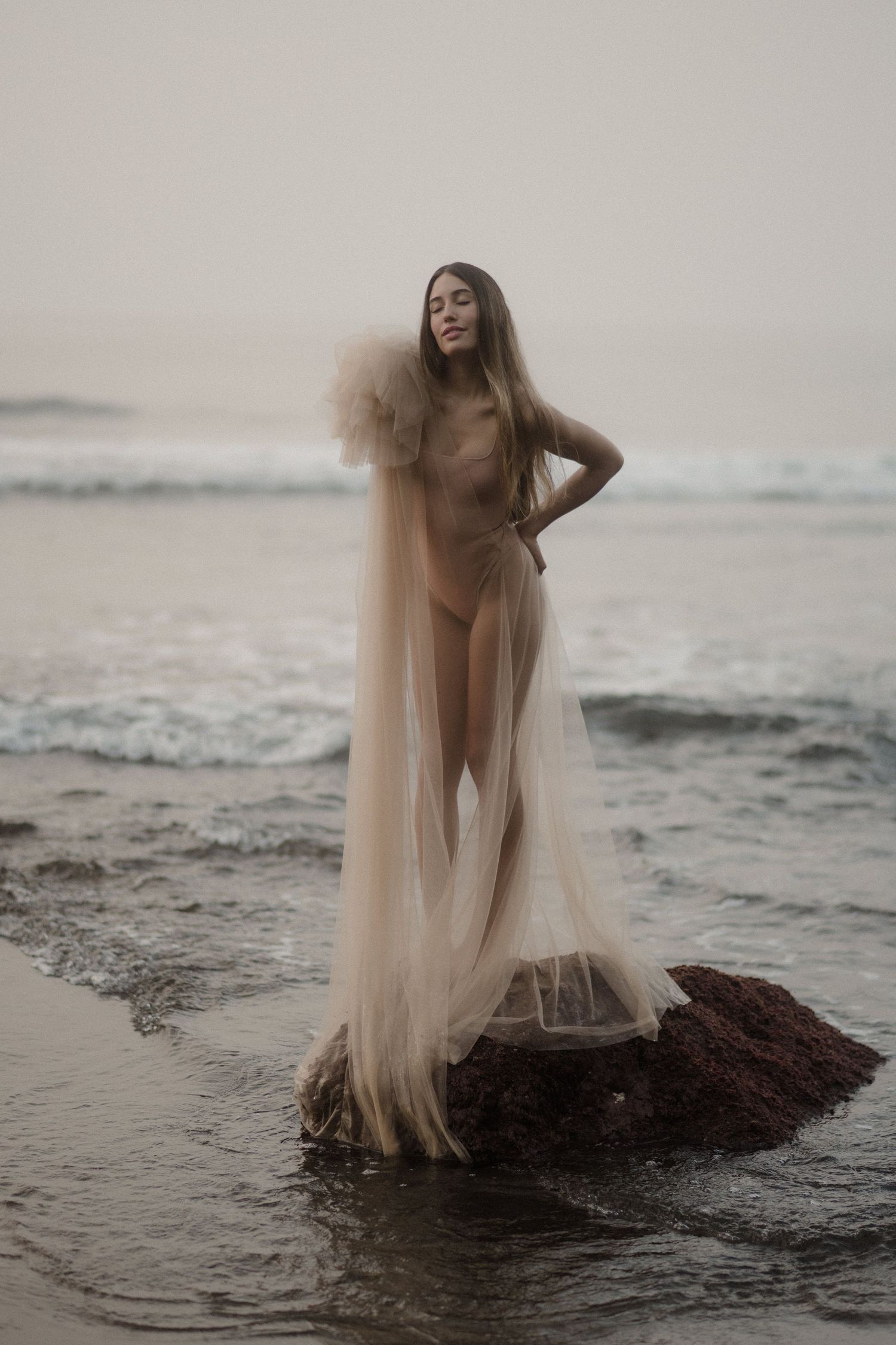 A figure draped in ethereal fabric poses gracefully on dark rocks by the ocean at sunset.