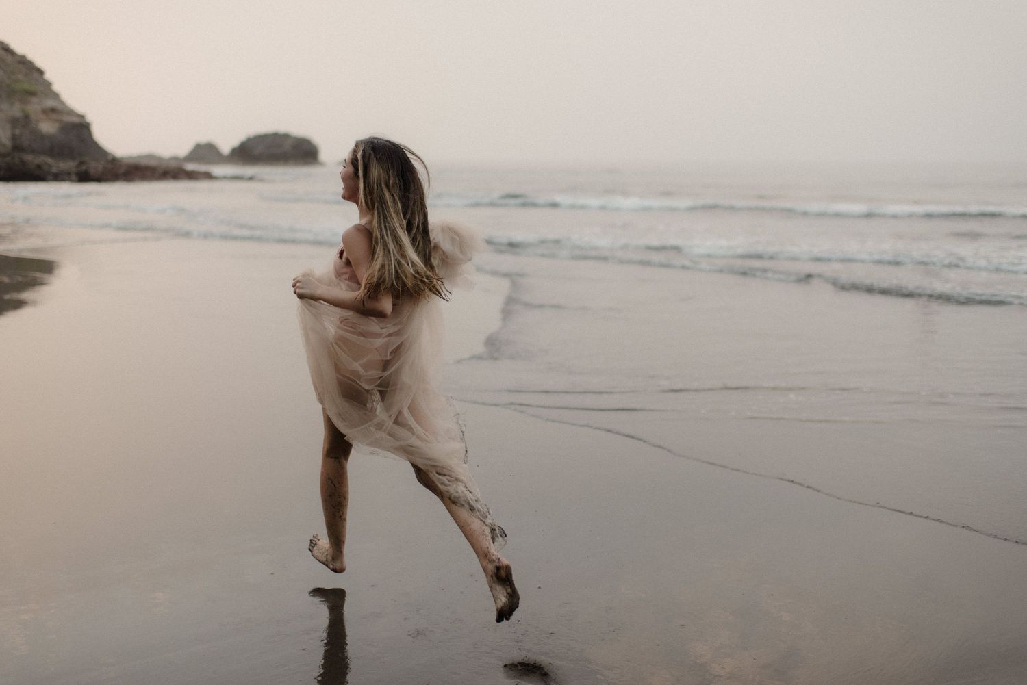 Ethereal figure in flowing dress walks gracefully along misty beach at sunset.