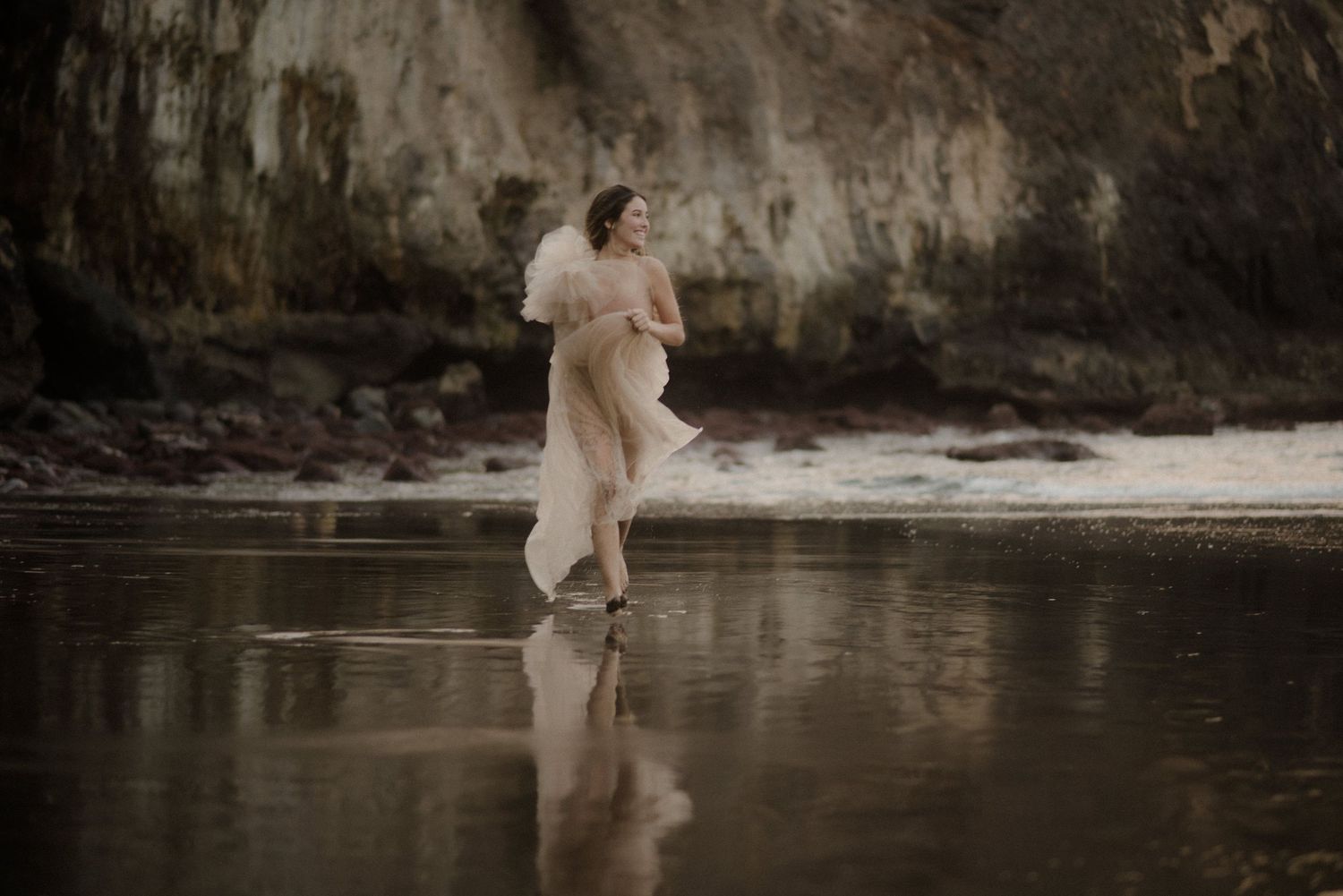 Artistic photoshoot in water shows figure in flowy dress against dramatic rocky cliffs.