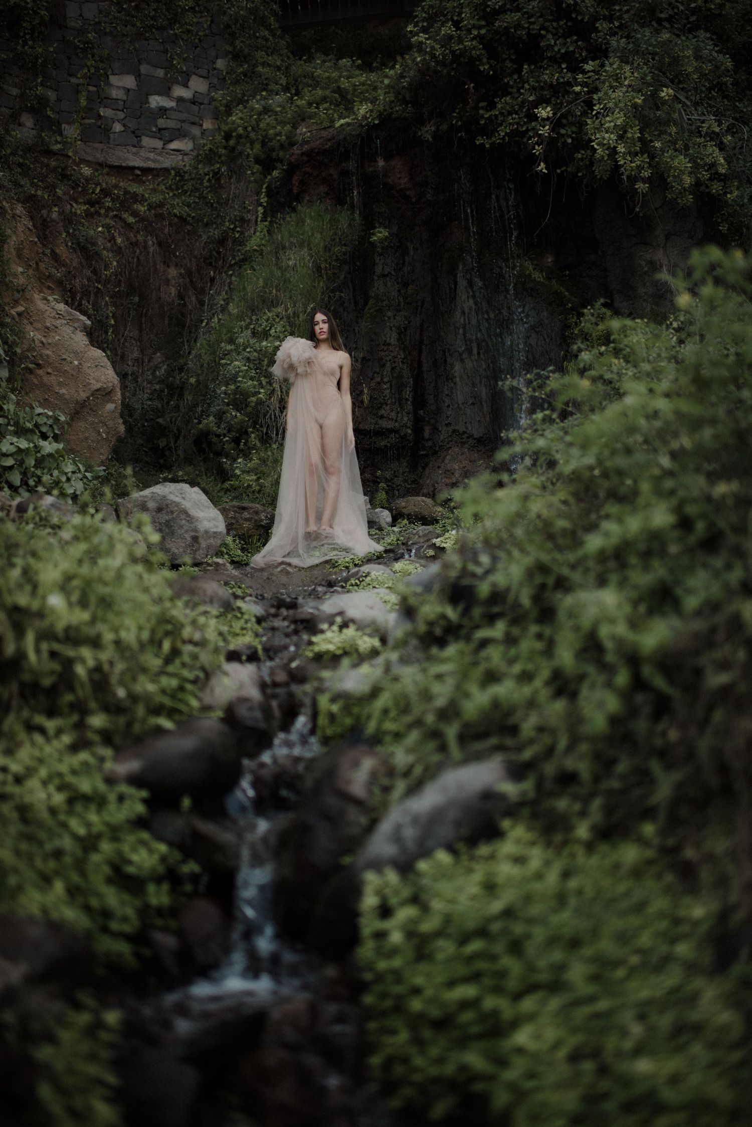 A dreamy portrait in a flowy cream dress standing by a mossy cliff wall near a cascading stream in moody lighting.