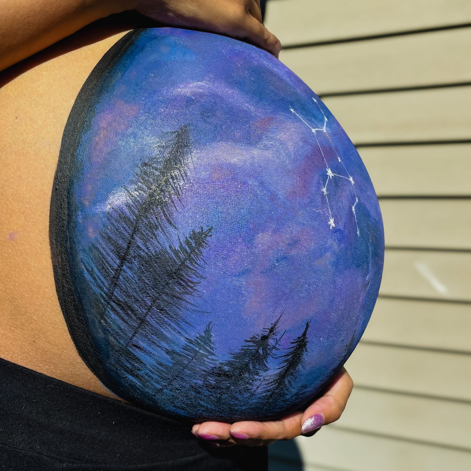 Painted landscape scene on pregnant belly showing starry night sky and forest silhouettes.